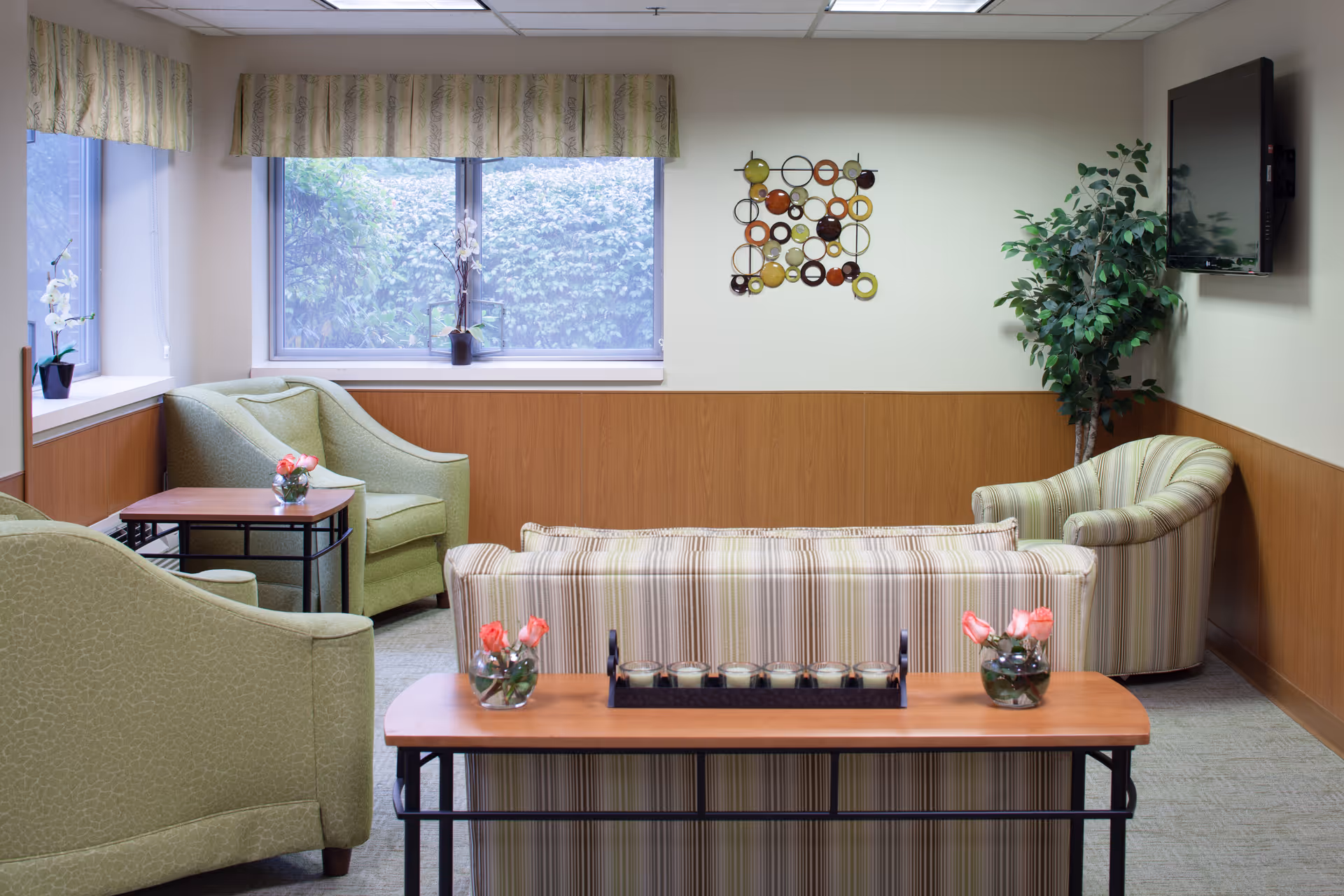 A cozy living room area with a striped sofa facing away, two green armchairs, a small wooden table with pink flowers in glass vases, a decorative wall art piece with colorful circular patterns, a potted plant in the corner, and a flat-screen TV mounted on the wall. Large windows with patterned valances let in natural light and show greenery outside.