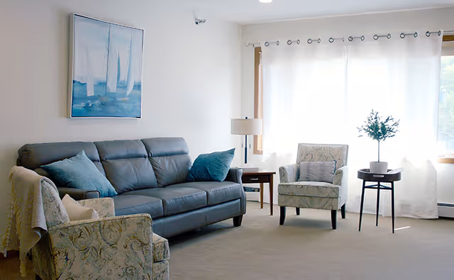 Bright living room with a gray sofa, two patterned armchairs, side tables, a lamp, and a large curtained window.