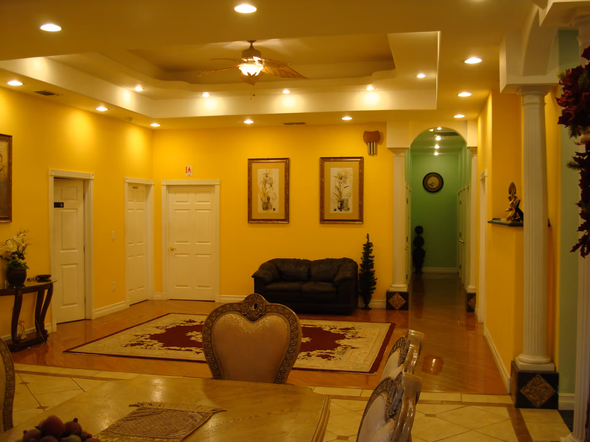 Interior view of a senior living facility room with yellow walls, a ceiling fan with lights, recessed ceiling lights, a dark leather couch, two framed floral pictures on the wall, a decorative rug on the floor, and a dining table with ornate chairs in the foreground. There is a hallway with green walls and decorative columns on the right side.