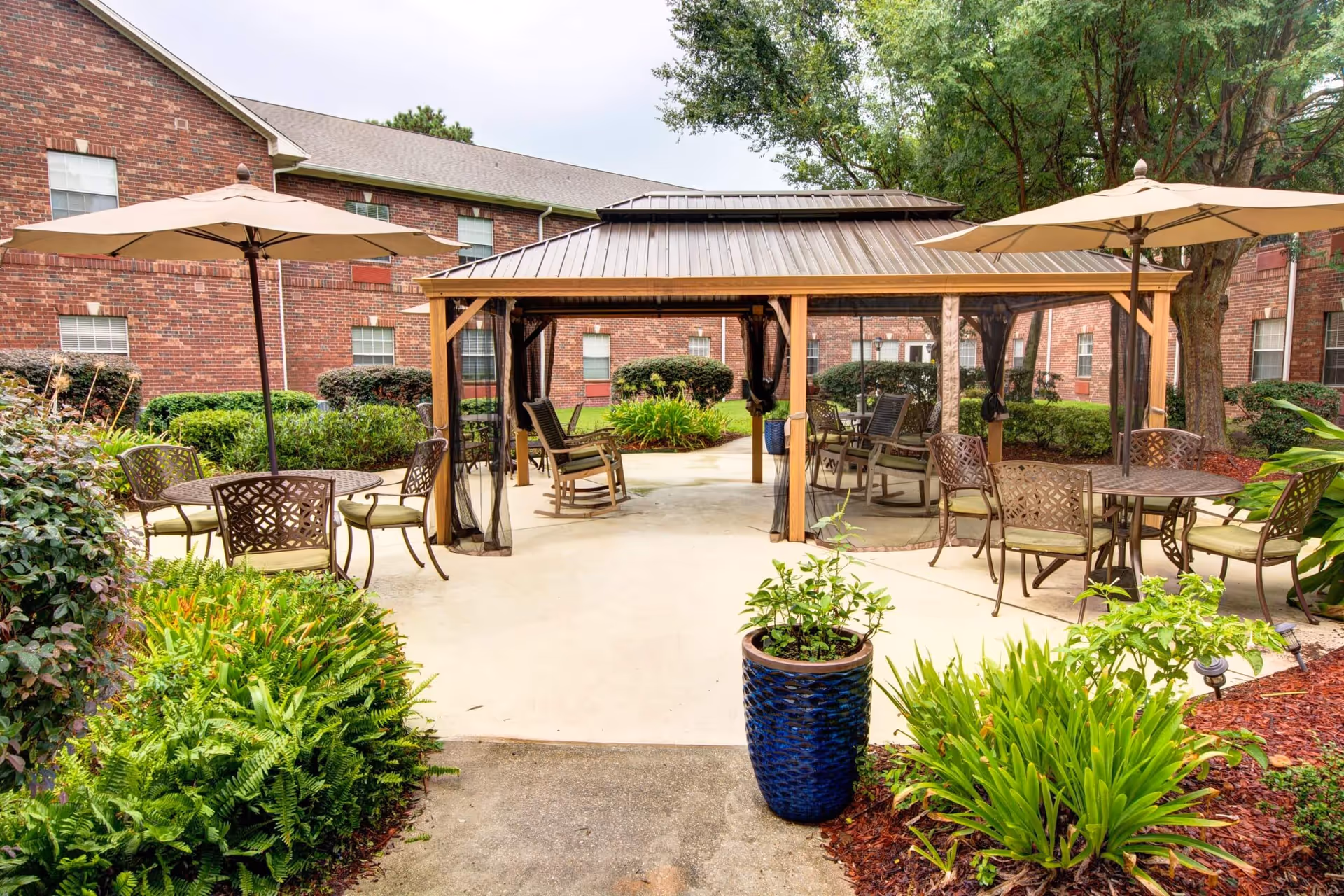 Outdoor patio area at Laketown Village featuring a covered gazebo with rocking chairs inside, surrounded by tables with umbrellas and metal chairs. The area is landscaped with green plants and shrubs, and brick buildings are visible in the background.