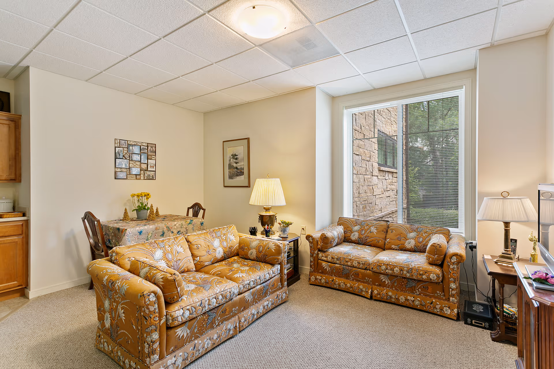 A cozy living room area with two matching floral-patterned sofas facing each other. There is a large window with blinds letting in natural light, a side table with a lamp between the sofas, and another side table with a lamp and decorative items near the window. In the background, there is a small dining table with a floral tablecloth and two chairs, along with framed pictures on the wall. The room has beige walls and carpeted flooring.