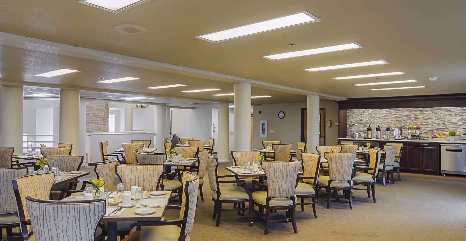 A spacious dining room with multiple tables and chairs arranged neatly. Each table is set with plates, cups, and utensils, and small flower arrangements are placed as centerpieces. The room has a neutral color scheme with beige walls and carpet, and there is a counter area in the back with coffee dispensers and fruit. The ceiling has recessed lighting, and there are several white columns throughout the space.