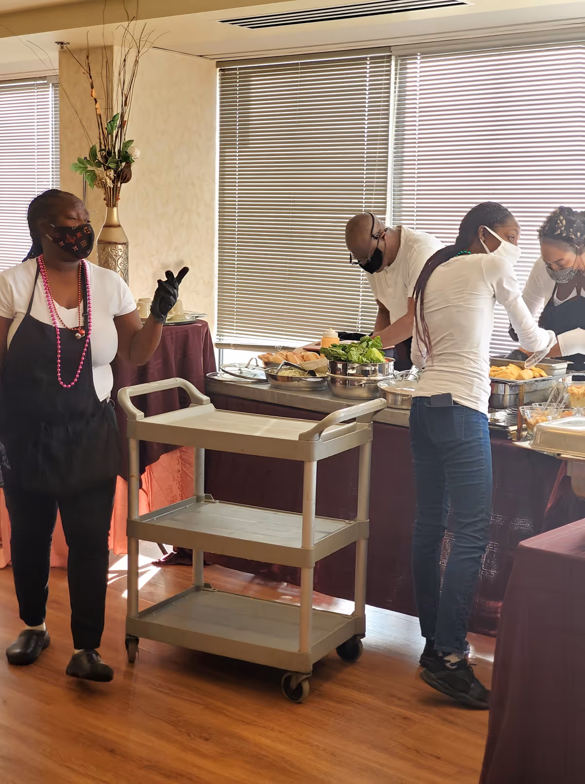Four people wearing masks and gloves are preparing and serving food from a buffet table in a room with wooden floors and large windows with blinds. One person is standing next to an empty three-tiered serving cart.