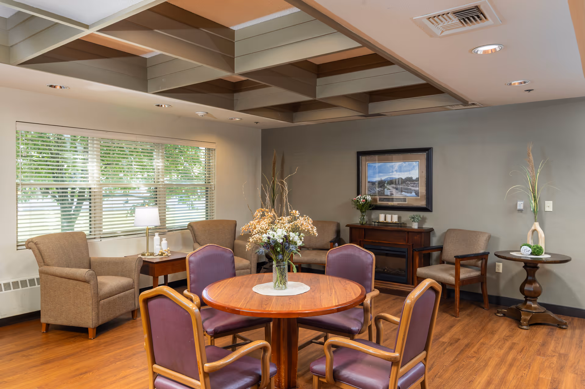Common lounge with a round wooden table and chairs, armchairs by a window, and decorative plants and artwork.