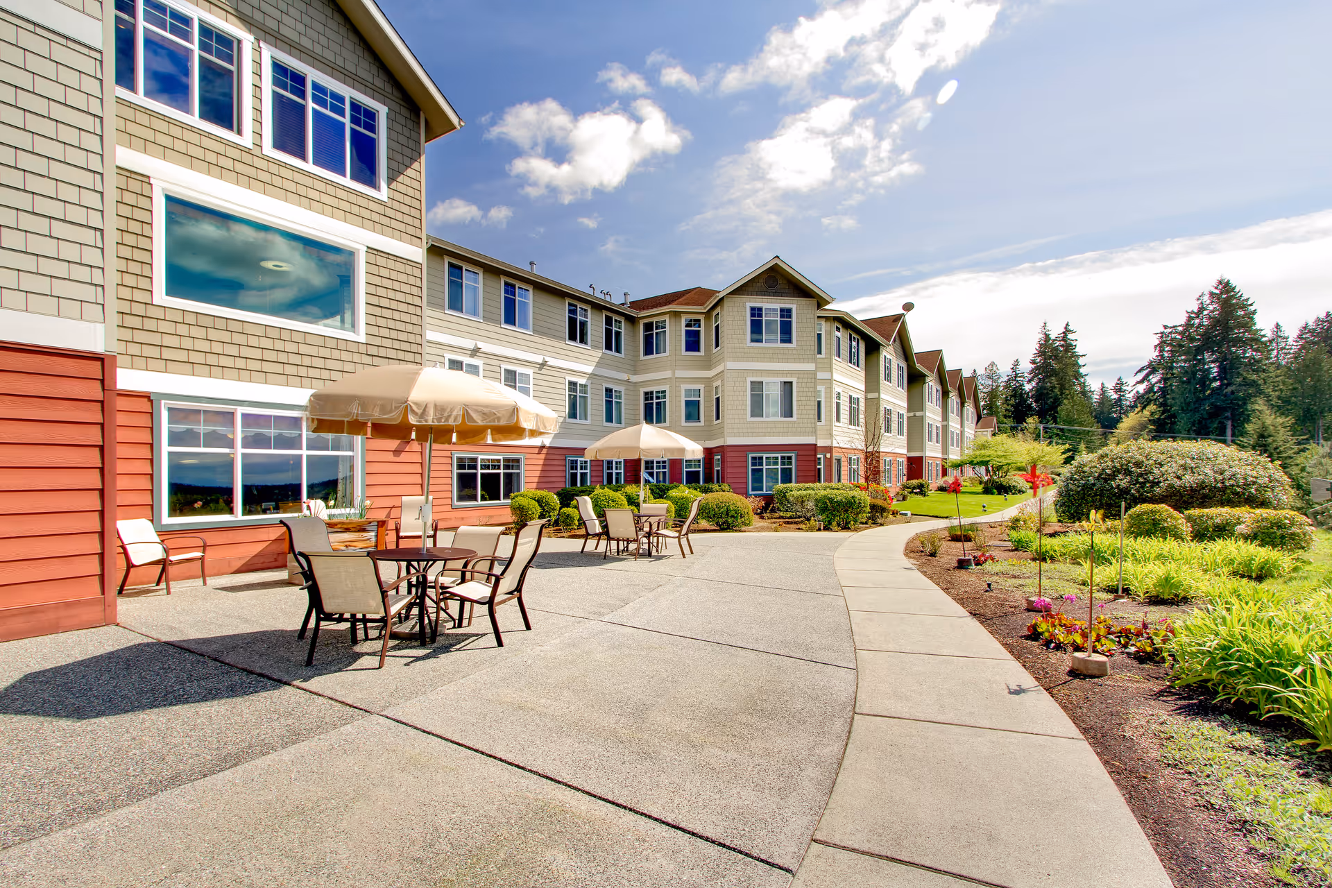 Outdoor patio area at Brookdale Fisher's Landing with tables, chairs, and umbrellas on a concrete surface next to a multi-story residential building. The area is surrounded by landscaped greenery, bushes, and trees under a partly cloudy sky.
