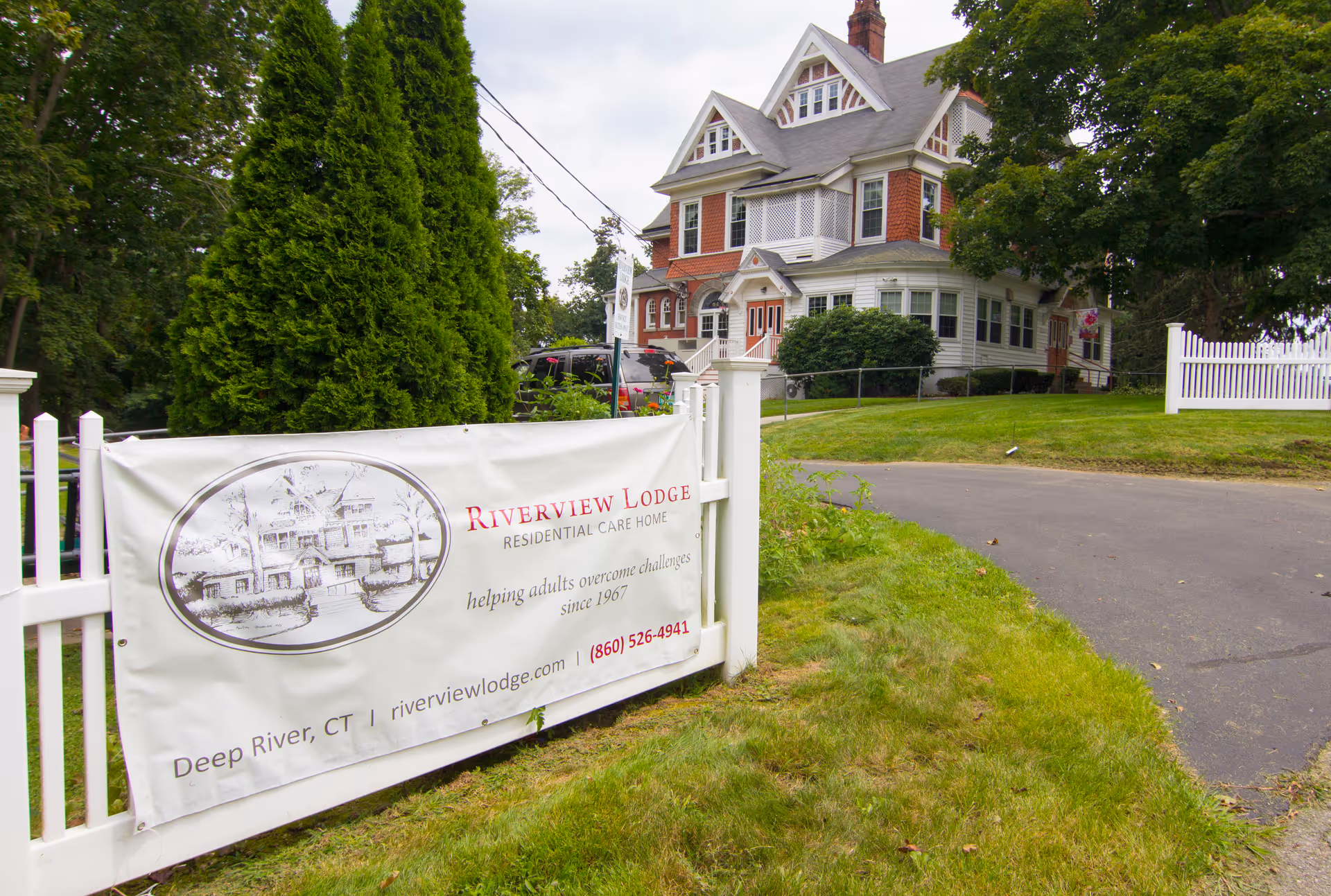 Front exterior of a large Victorian-style house with a Riverview Lodge Residential Care Home banner on a white fence.