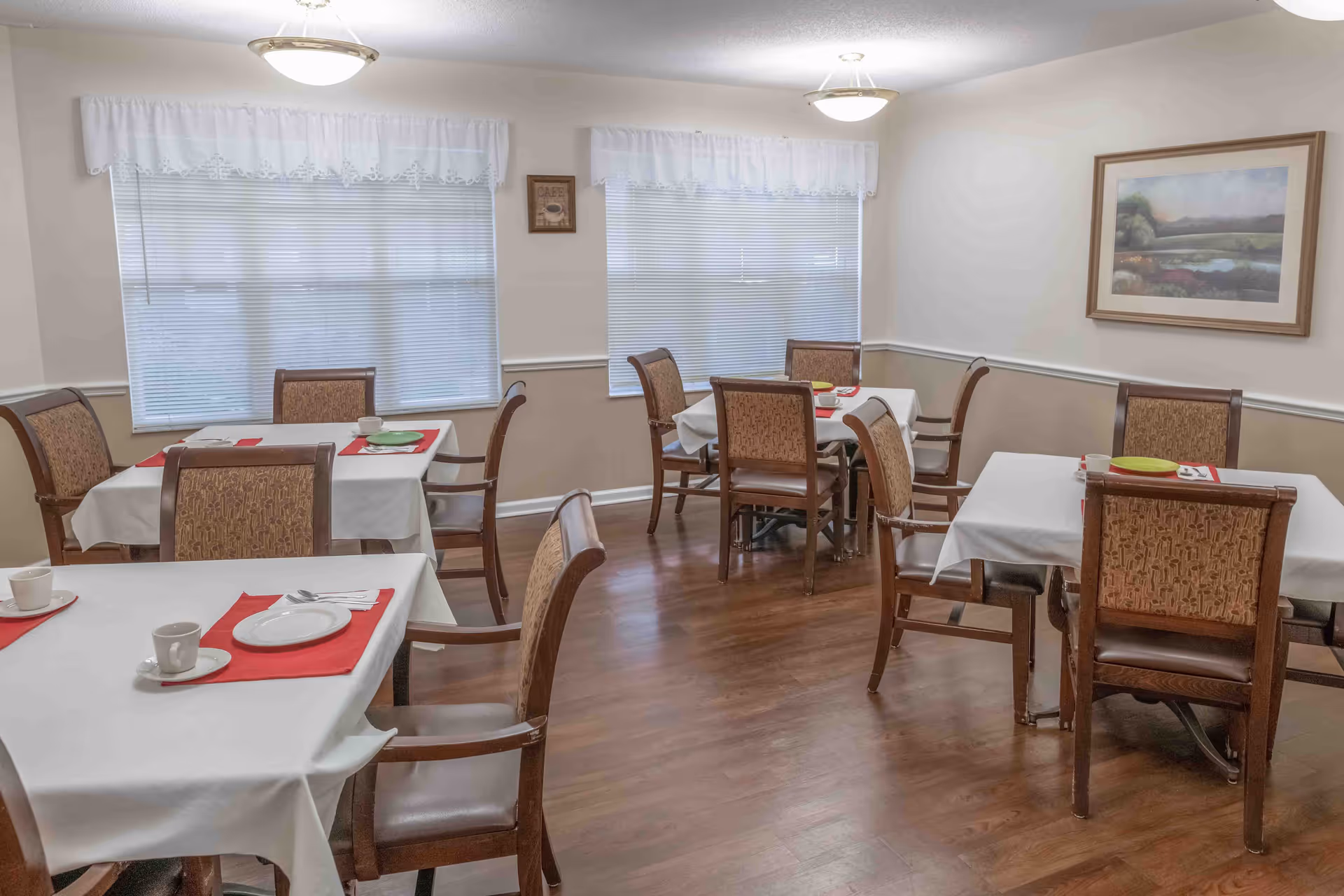 A dining room with four tables covered in white tablecloths and red placemats. Each table has wooden chairs with patterned upholstery. The room has two windows with white blinds and lace valances, a framed landscape painting on the wall, and wooden flooring.