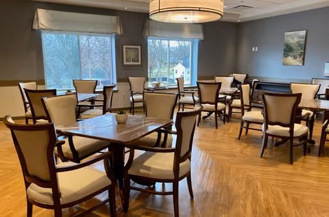 A well-lit dining room with multiple wooden tables and cushioned chairs arranged neatly. Large windows let in natural light and offer a view of trees and a small building outside. The room features wooden flooring, gray walls, framed artwork, and a large circular ceiling light fixture.