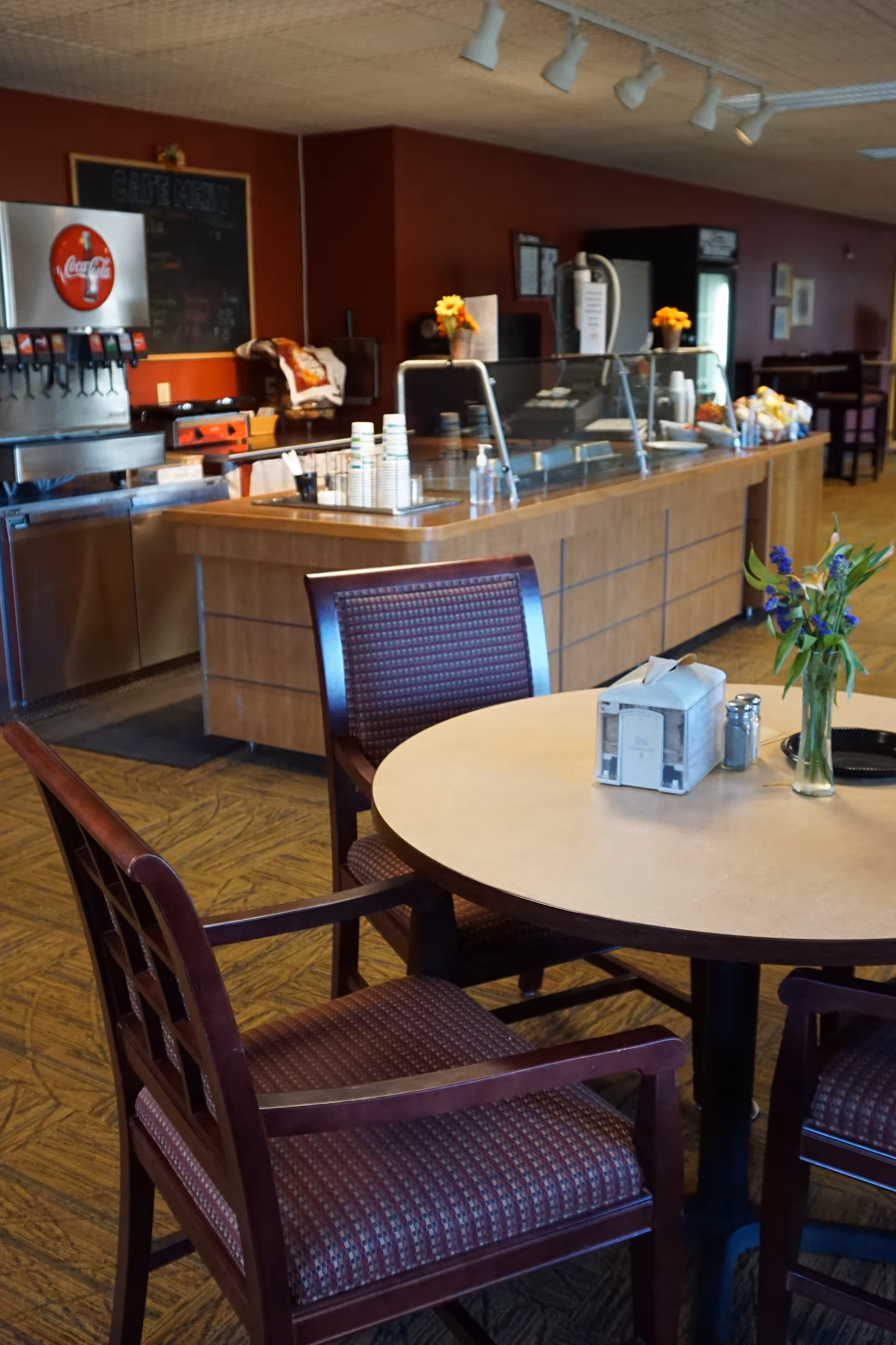 Round dining table with chairs in the foreground and a self-serve cafeteria counter with a soda dispenser and flowers in the background.