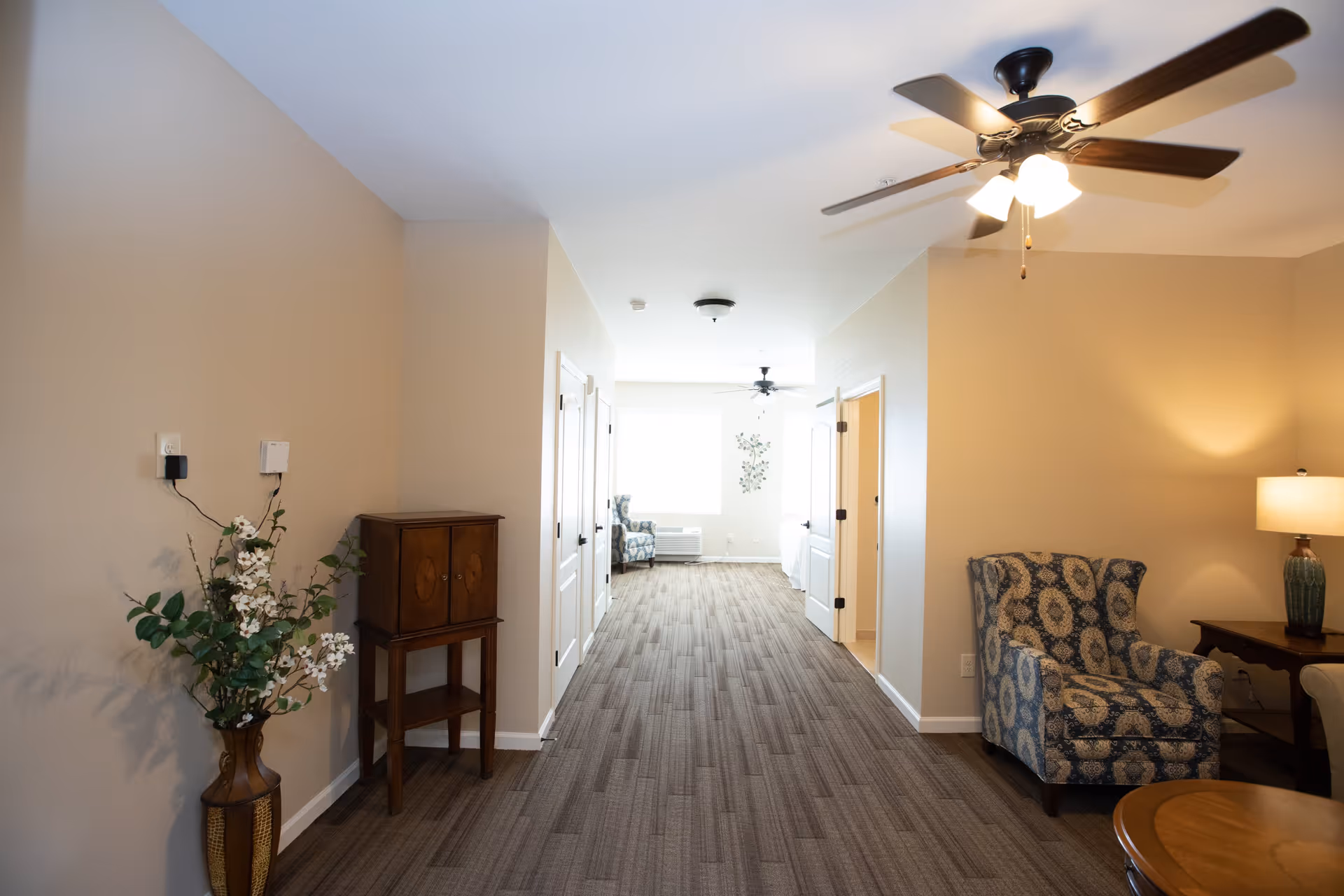 Well-lit interior hallway and sitting area with patterned armchairs, ceiling fans, a table lamp, and decorative plants.
