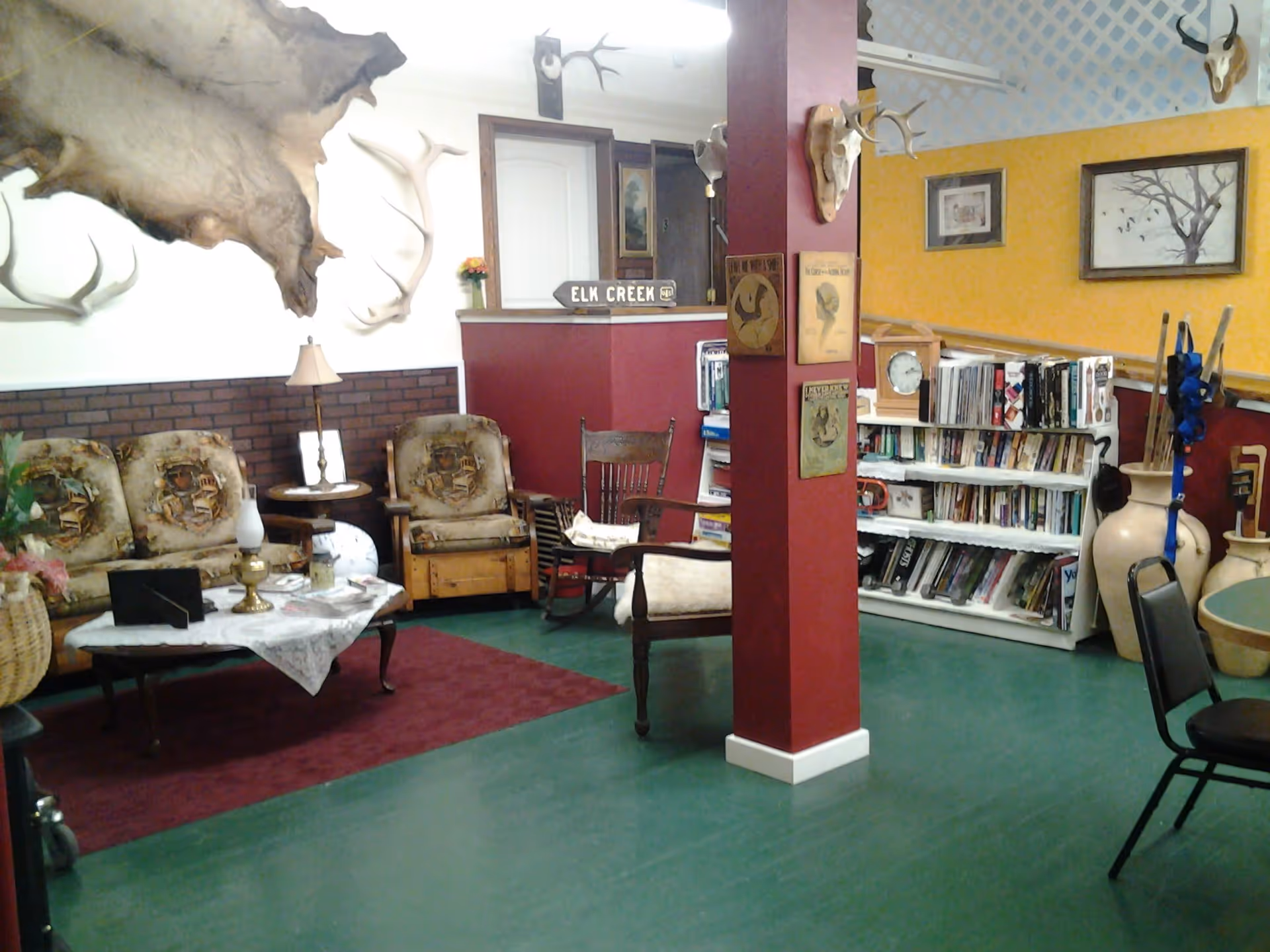 A cozy common area with vintage-style furniture including a floral-patterned sofa and armchair, a wooden rocking chair, and a small table with a lace tablecloth and lamps. The walls are decorated with mounted antlers, animal skulls, and framed pictures. A bookshelf filled with books is visible in the background, along with a red pillar adorned with vintage posters. The floor is green with a maroon rug under the seating area.