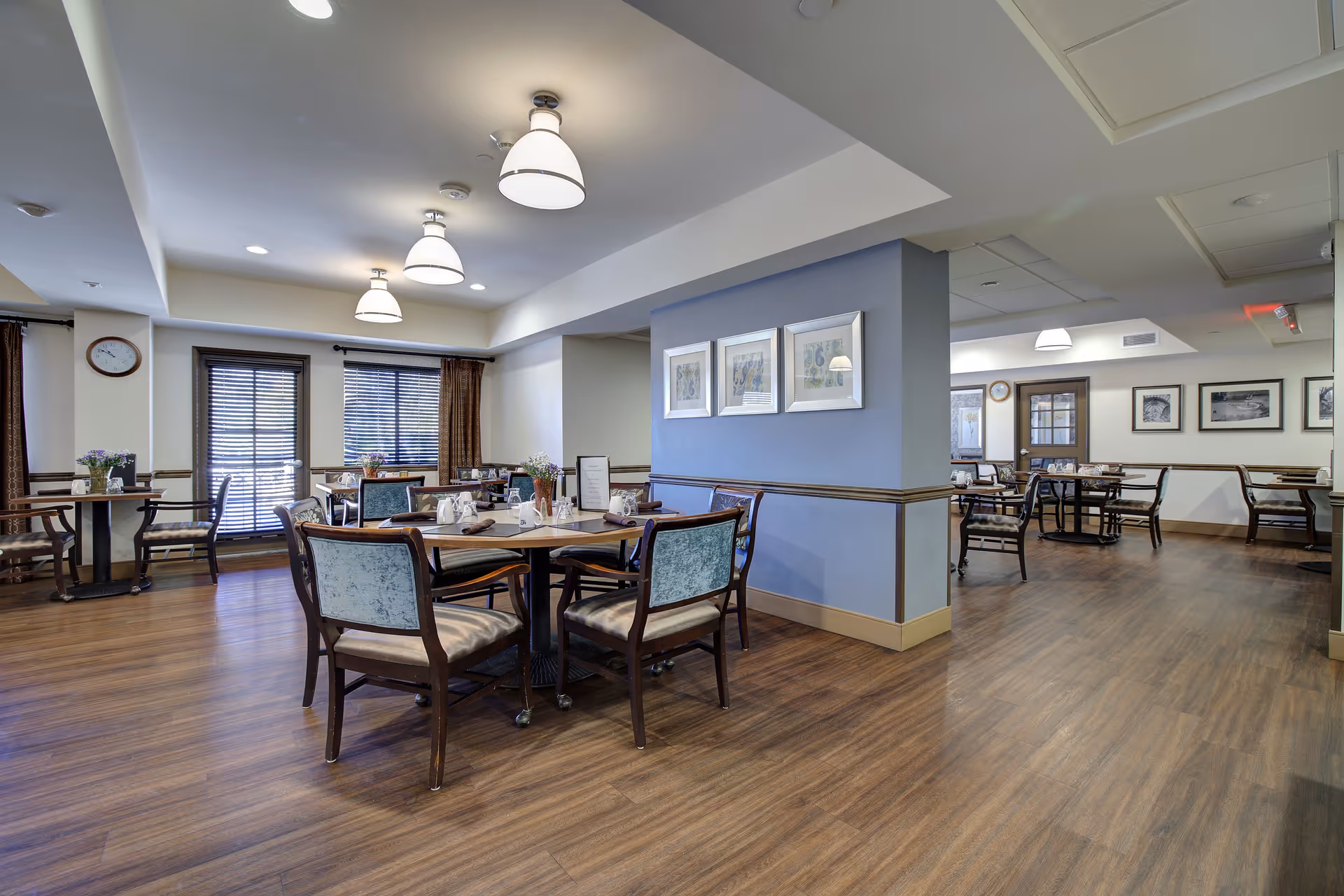 A spacious senior living dining room with multiple wooden tables and chairs arranged neatly. The room features wood flooring, soft lighting from ceiling fixtures, framed artwork on the walls, and large windows with blinds and curtains allowing natural light to enter.