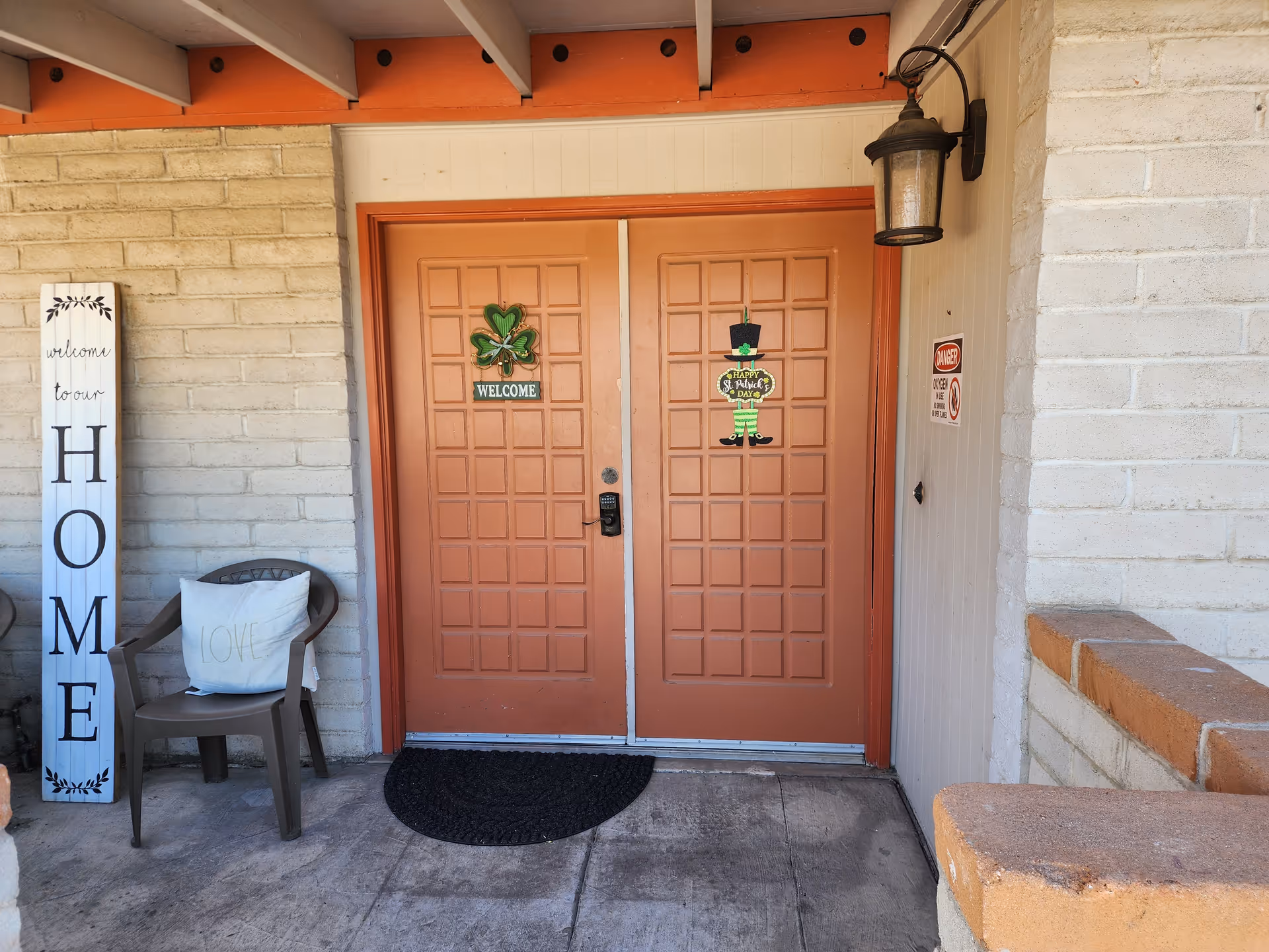 Front entrance of a building with double orange doors decorated with a shamrock welcome sign and a St. Patrick's Day decoration. To the left of the doors is a plastic chair with a white pillow that says 'LOVE' and a tall vertical wooden sign that reads 'welcome to our HOME'. The walls are made of light-colored bricks and there is a black outdoor lantern mounted on the wall to the right of the doors.