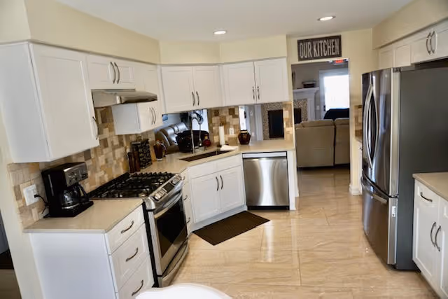 Bright modern kitchen with white cabinetry, stainless steel appliances, tiled backsplash, and a view into an adjoining living area.