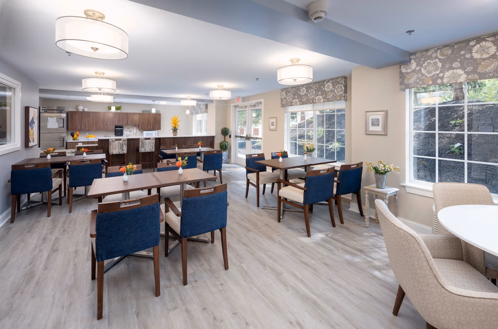 A bright and spacious dining area in a senior living facility featuring multiple wooden tables with blue and beige cushioned chairs. The room has large windows with floral valances allowing natural light to fill the space. There is a kitchen area in the background with wooden cabinets, a refrigerator, microwave, and a counter with bar stools. The floor is light-colored wood, and the ceiling has modern round light fixtures. Small flower arrangements are placed on each table.