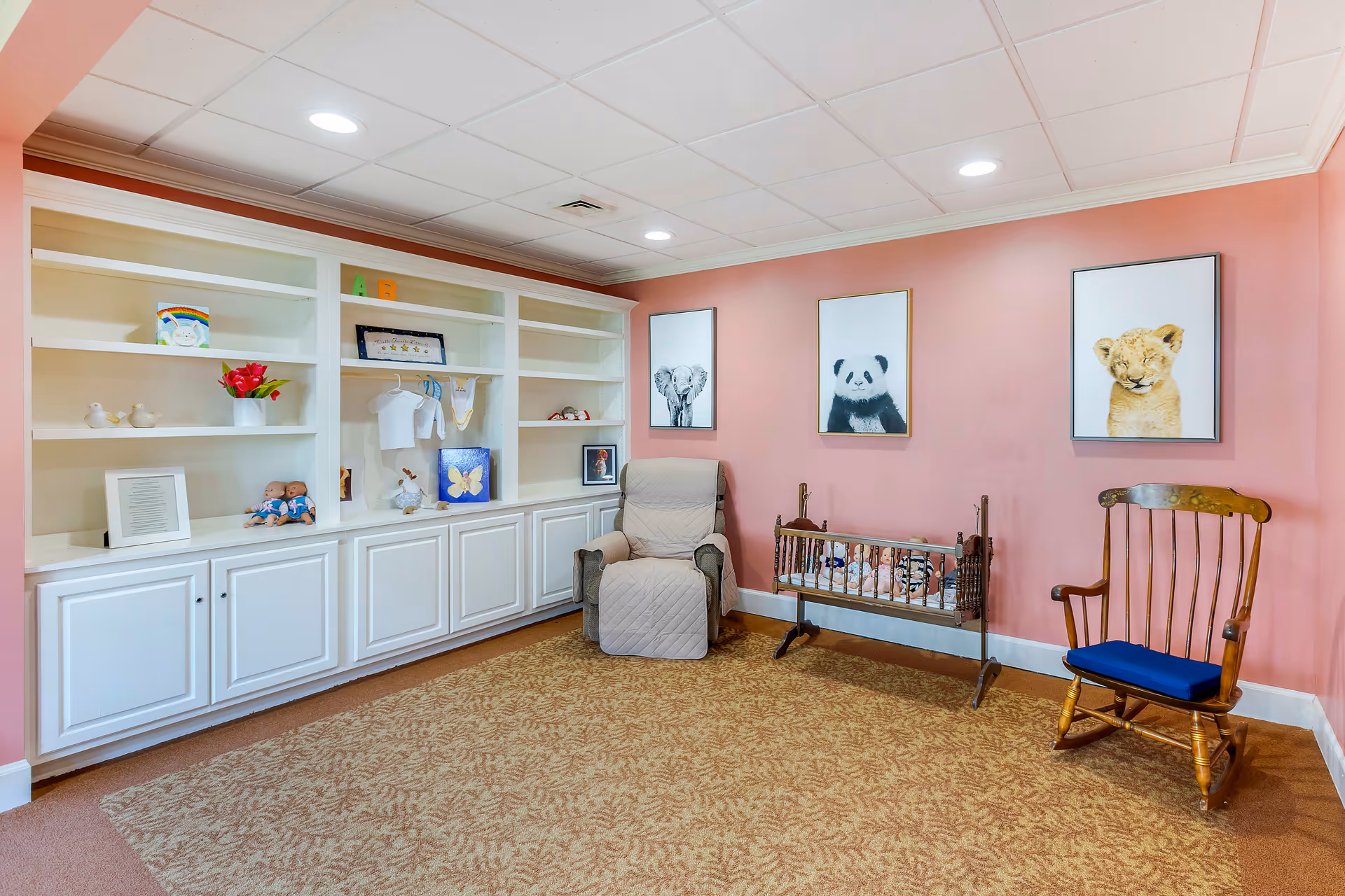 A cozy room with pink walls featuring three framed animal pictures of an elephant, a panda, and a lion cub. The room has a beige patterned carpet, a cushioned recliner chair, a wooden rocking chair with a blue cushion, and a vintage wooden cradle with stuffed animals inside. There is a large white built-in shelving unit with various decorative items including dolls, a framed picture, and a small bouquet of flowers.
