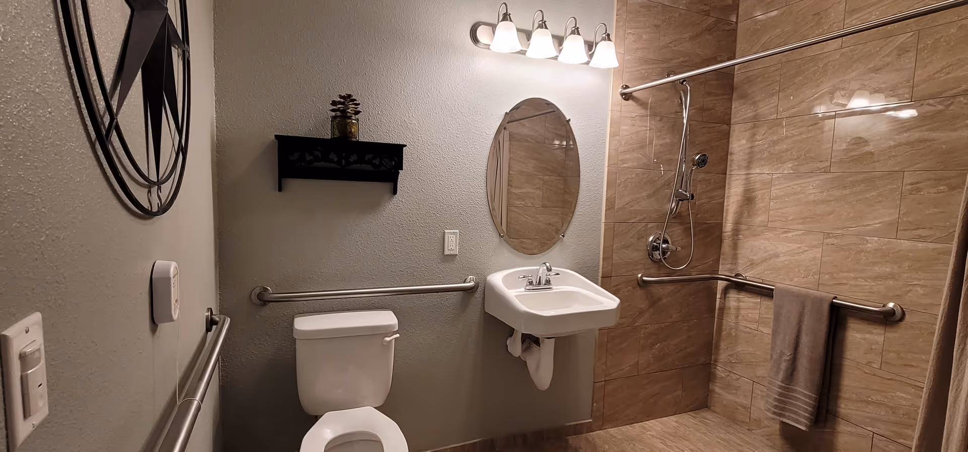 A bathroom with a toilet, wall-mounted sink, oval mirror, and a walk-in shower with a handheld showerhead. The walls are tiled in the shower area and painted light gray elsewhere. There are grab bars near the toilet and in the shower, a towel hanging on one grab bar, a decorative shelf with a small plant above the toilet, and a round wall decoration on the left wall. The lighting fixture above the mirror has five lights.