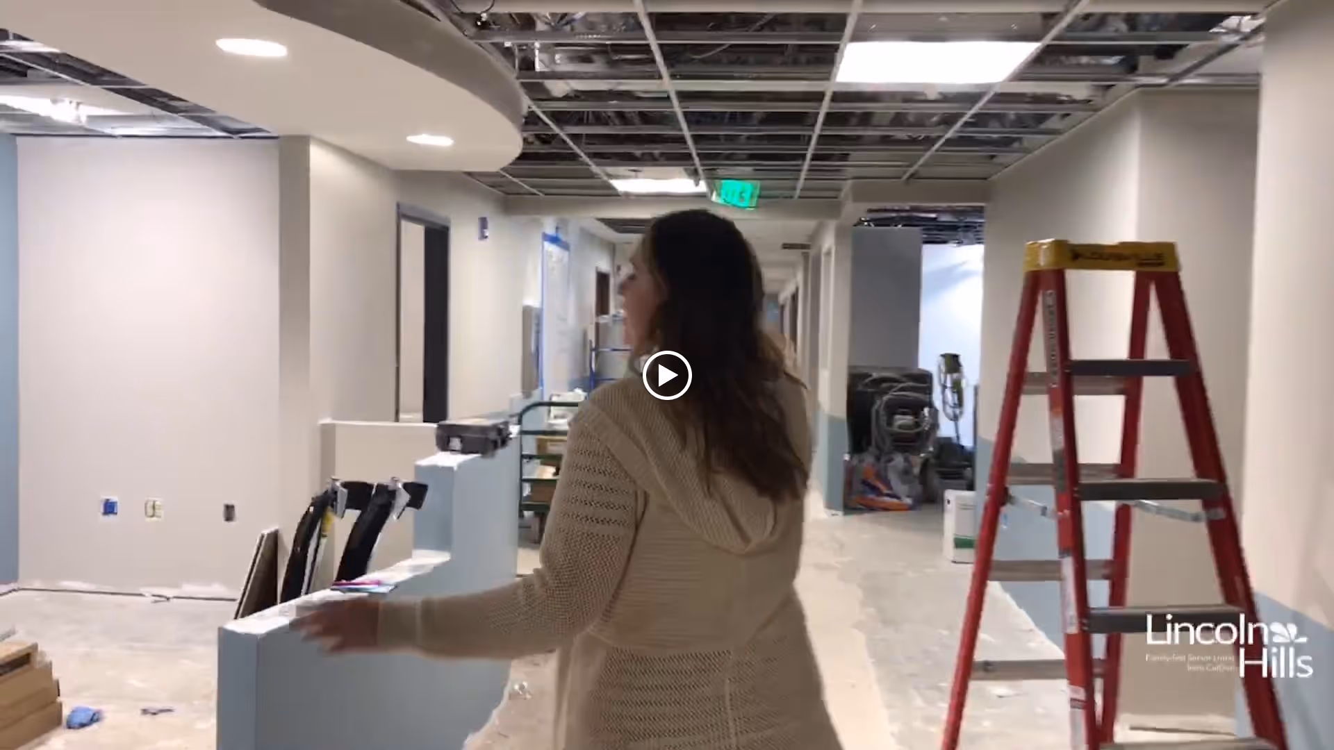 A woman with long brown hair wearing a beige knitted sweater walks through a hallway under construction with exposed ceiling and unfinished walls. A red ladder and construction materials are visible along the hallway. The Lincoln Hills logo is visible in the bottom right corner.