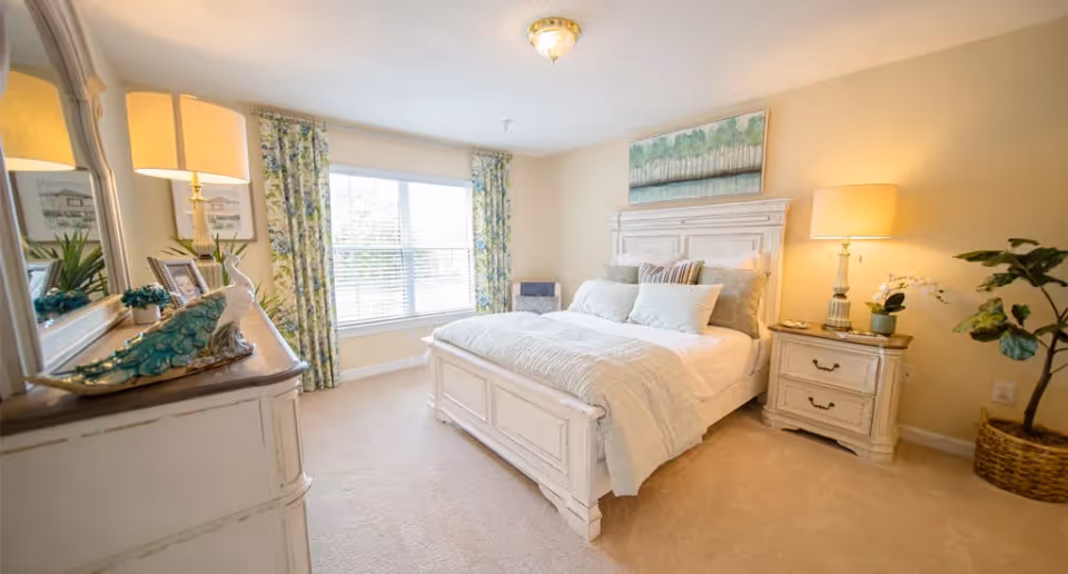 A bright and cozy bedroom with a large window covered by floral curtains. The room features a white wooden bed with multiple pillows and a light blue blanket. On the right side, there is a matching white nightstand with a lamp and a small plant. On the left side, a white dresser with a large mirror holds decorative items including a peacock figurine and framed photos. The walls are painted a soft beige color, and a painting of trees hangs above the bed.
