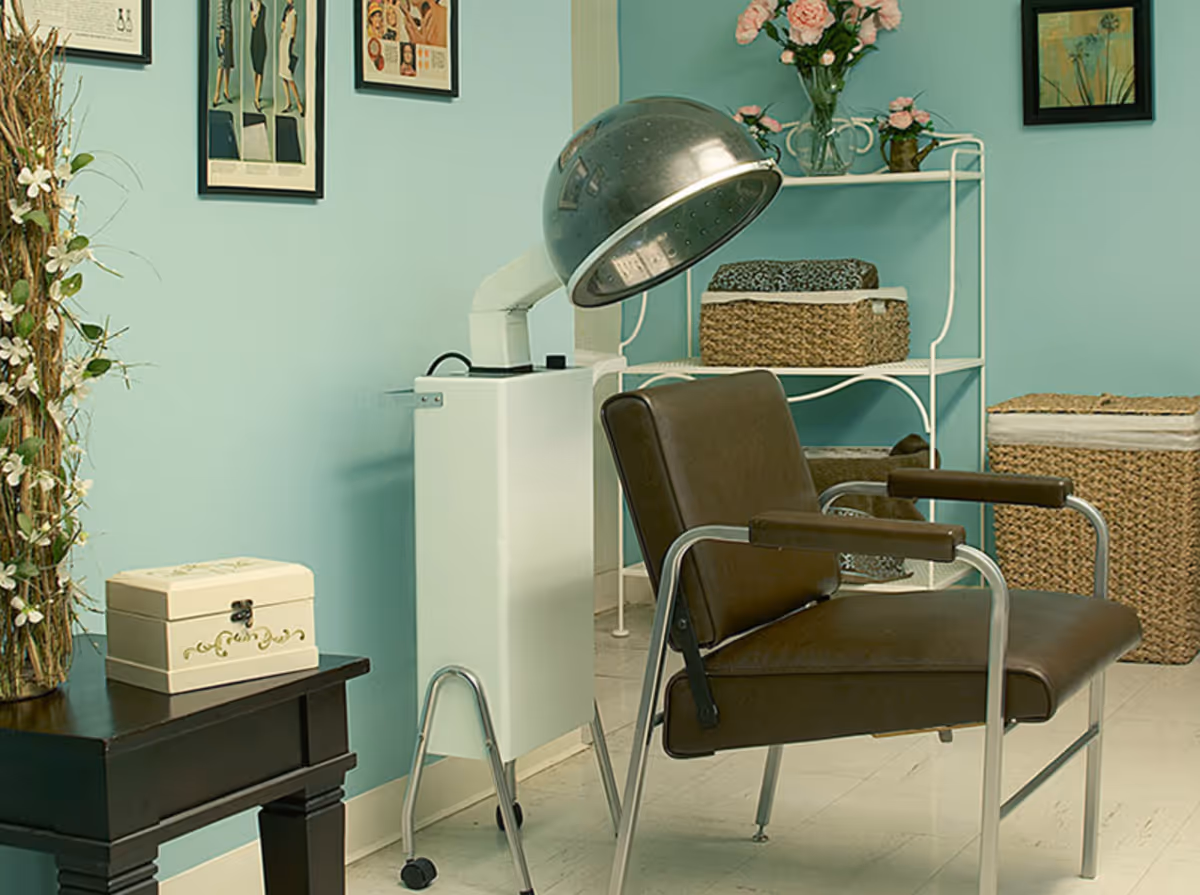 A vintage hair drying chair with a hooded hair dryer in a room with light blue walls. The room has a small black table with a decorative box, a white metal shelf with woven baskets and a vase of pink flowers, and framed pictures on the wall.