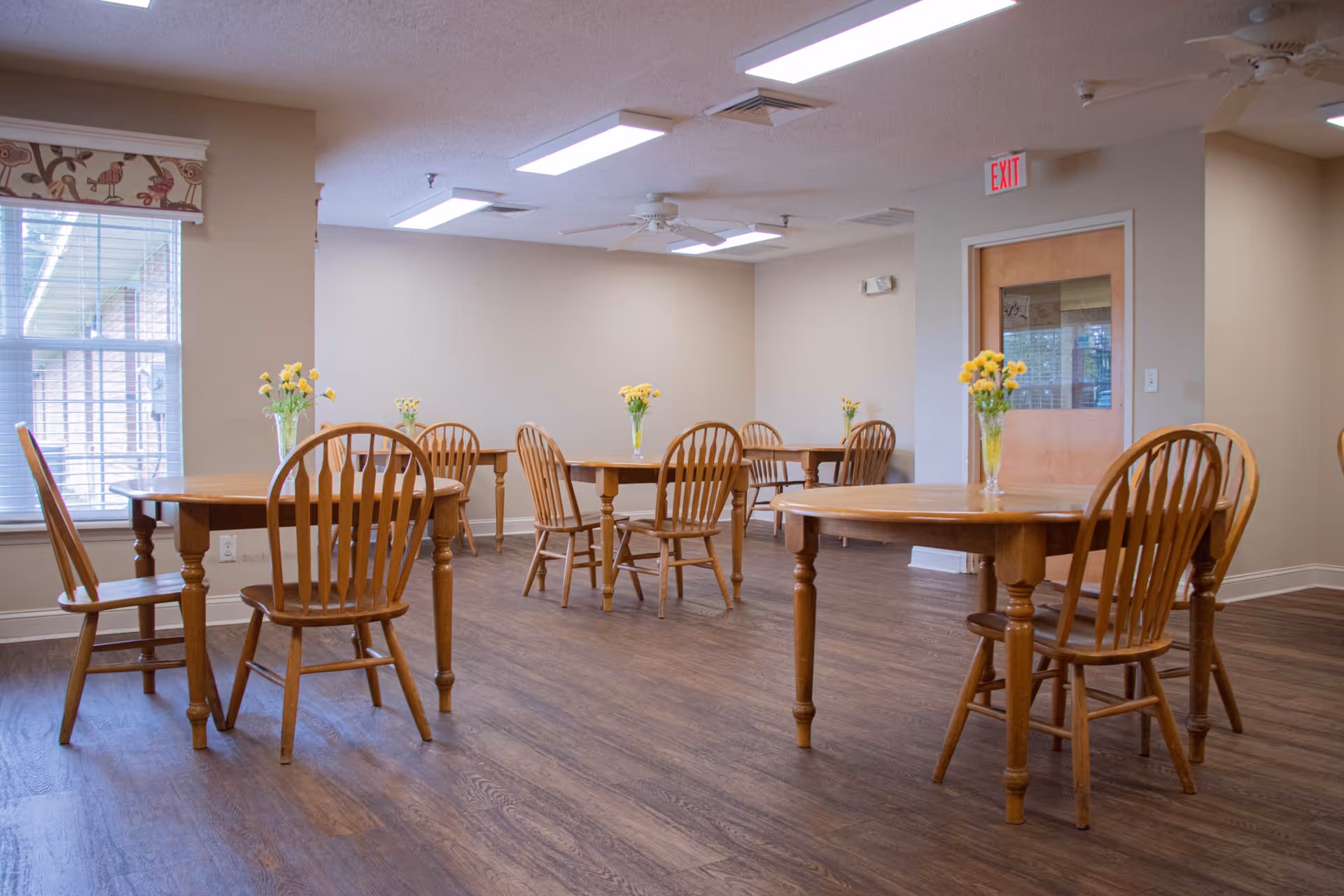 A dining room with several wooden tables and chairs arranged neatly. Each table has a vase with yellow flowers. The room has wood flooring, beige walls, a window with blinds and a patterned valance, and a door with a glass panel and an exit sign above it.