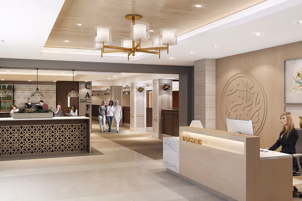 Bright modern lobby showing a welcome desk with a receptionist, decorative wall panel and chandelier, and people walking toward a café/reception area.