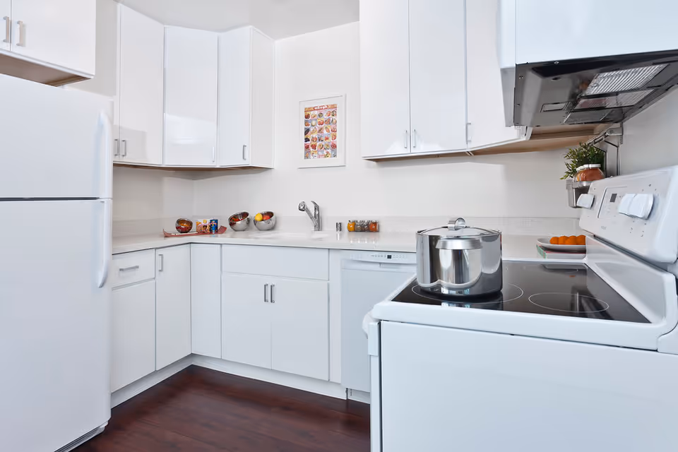 A modern kitchen with white cabinets, a white refrigerator, a white electric stove with a stainless steel pot on it, a dishwasher, and a sink. The countertop has bowls with fruit, small jars of spices, and a framed picture on the wall. The floor is dark wood.