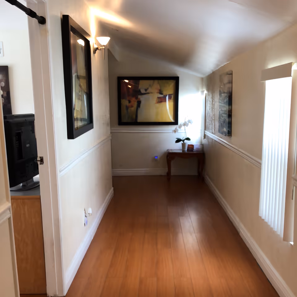 A narrow hallway with wooden flooring and cream-colored walls. There are framed abstract paintings on both walls, a small wooden table with a white orchid plant at the end of the hallway, and a wall-mounted light fixture on the left wall. A window with vertical blinds is on the right side, allowing natural light to enter.