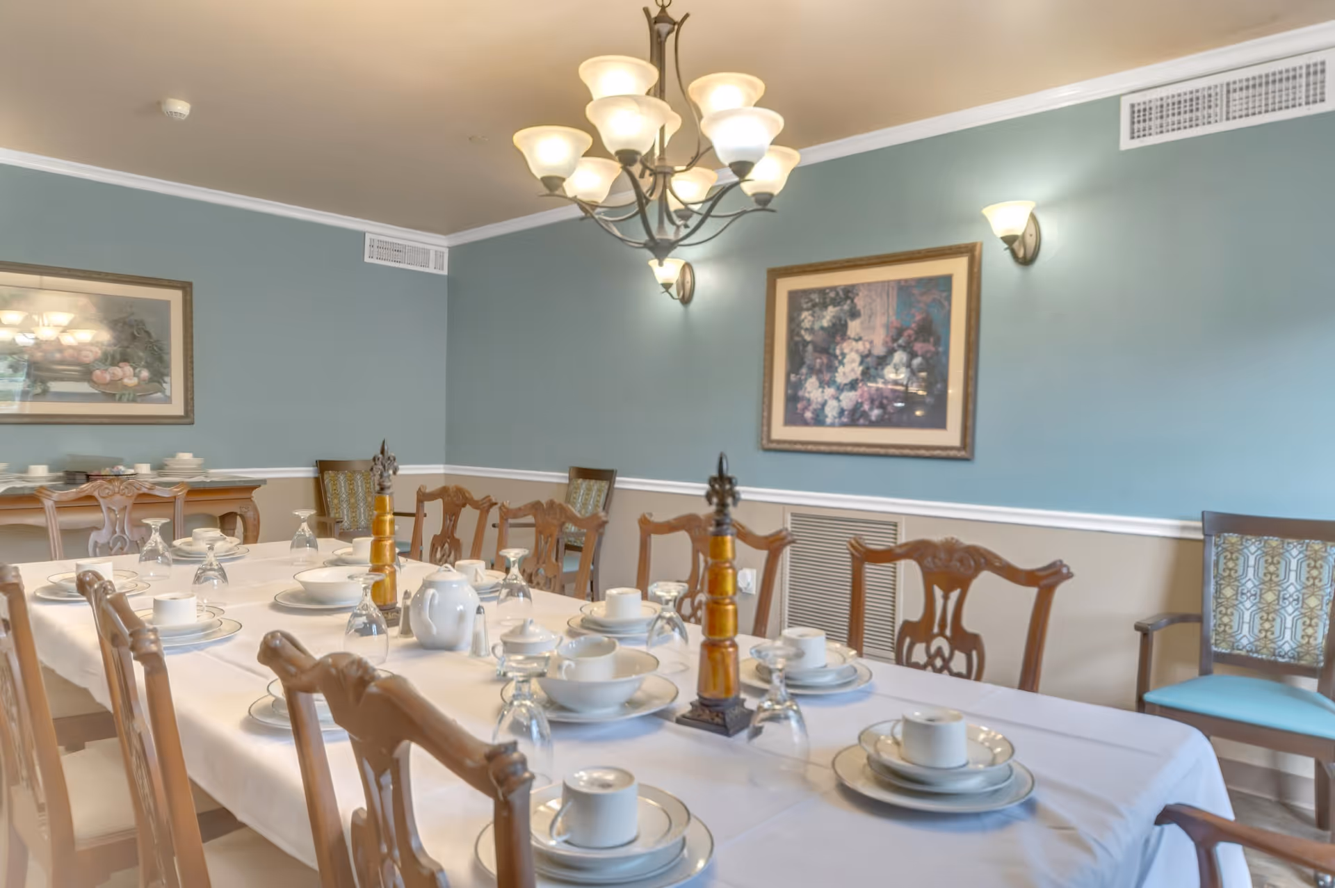 Formal dining room with a long table set with white dishes and teapots, wooden chairs, a chandelier, and framed artwork on blue-green walls.
