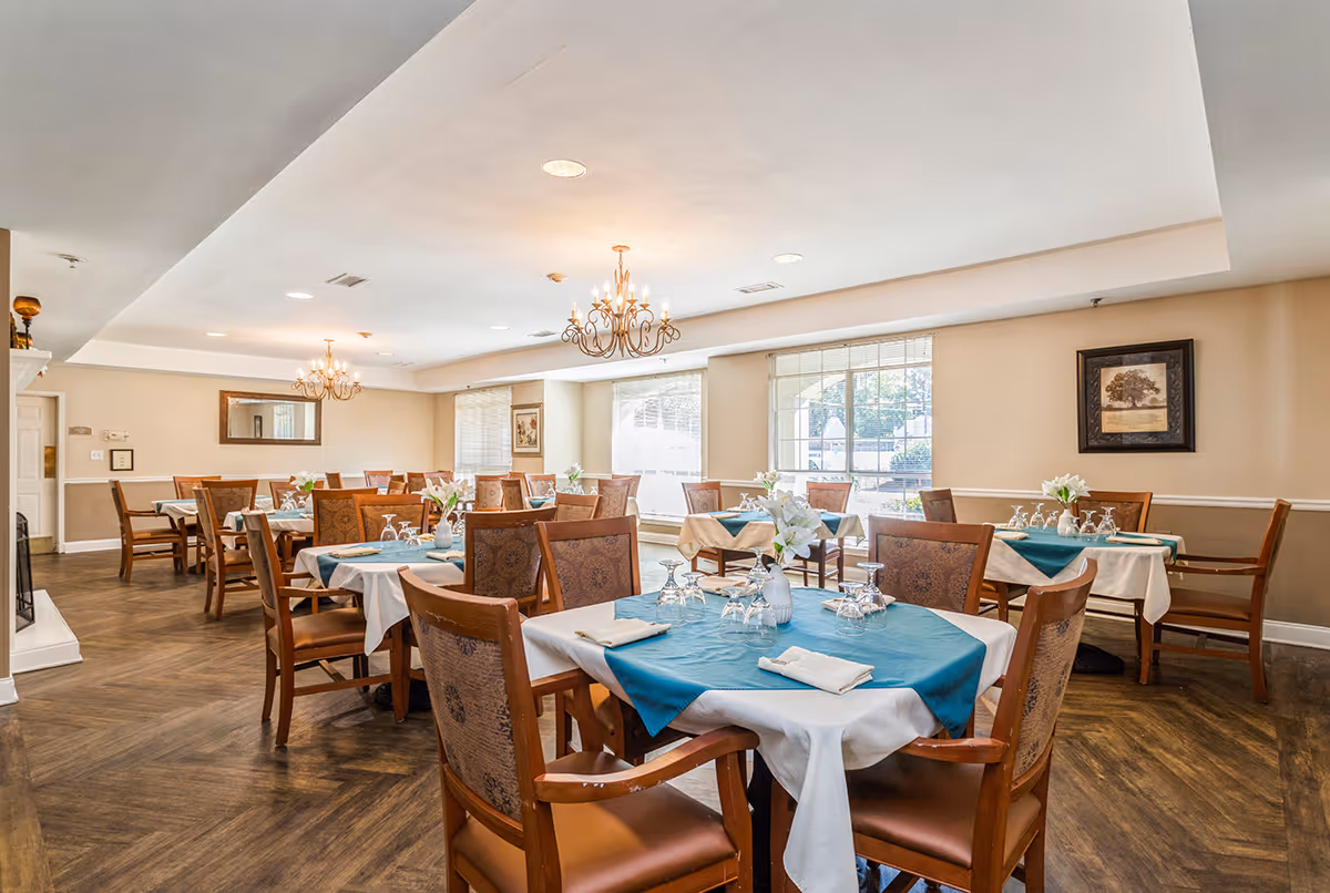 A bright and elegant dining room with multiple wooden tables and chairs. Each table is covered with white tablecloths and teal table runners, set with folded napkins, glassware, and small vases of white flowers. The room features large windows allowing natural light to fill the space, beige walls with framed artwork, and two ornate chandeliers hanging from the ceiling. The floor has a wood-patterned finish.