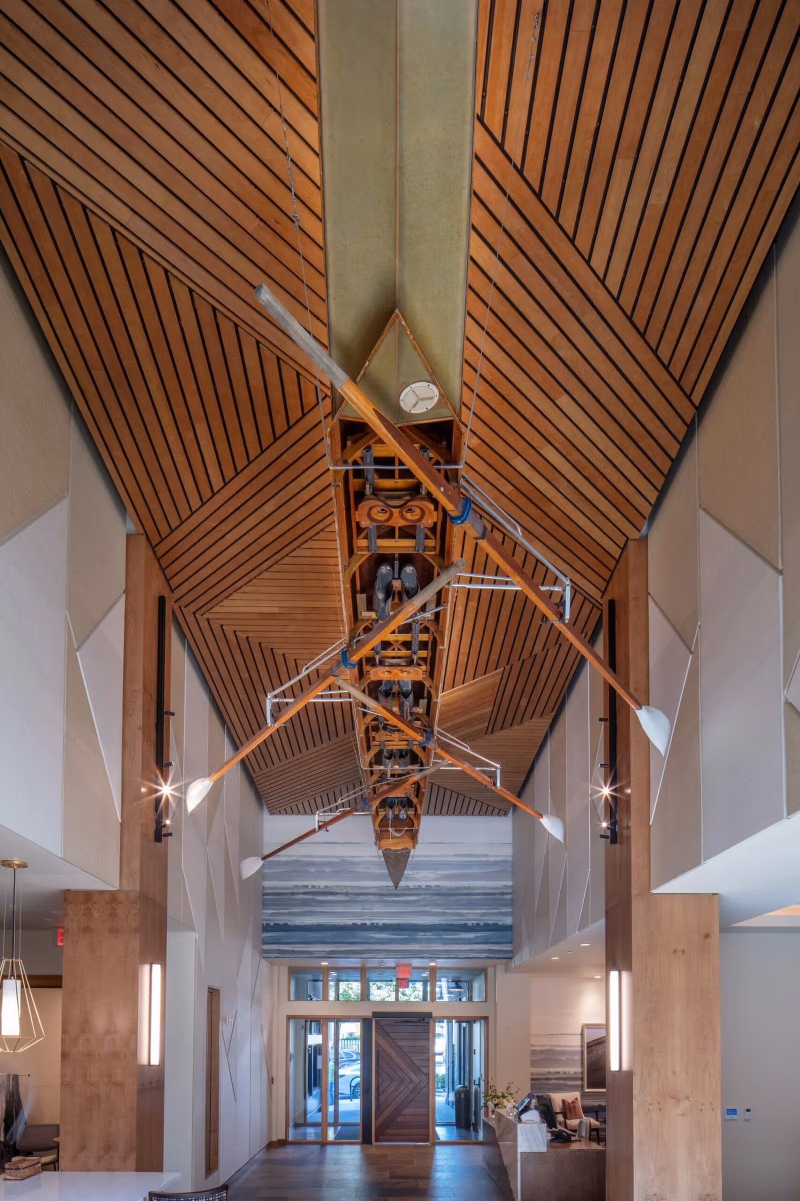 Lobby interior with a suspended wooden boat and oars beneath a patterned wooden ceiling, facing the entrance doors and reception area.