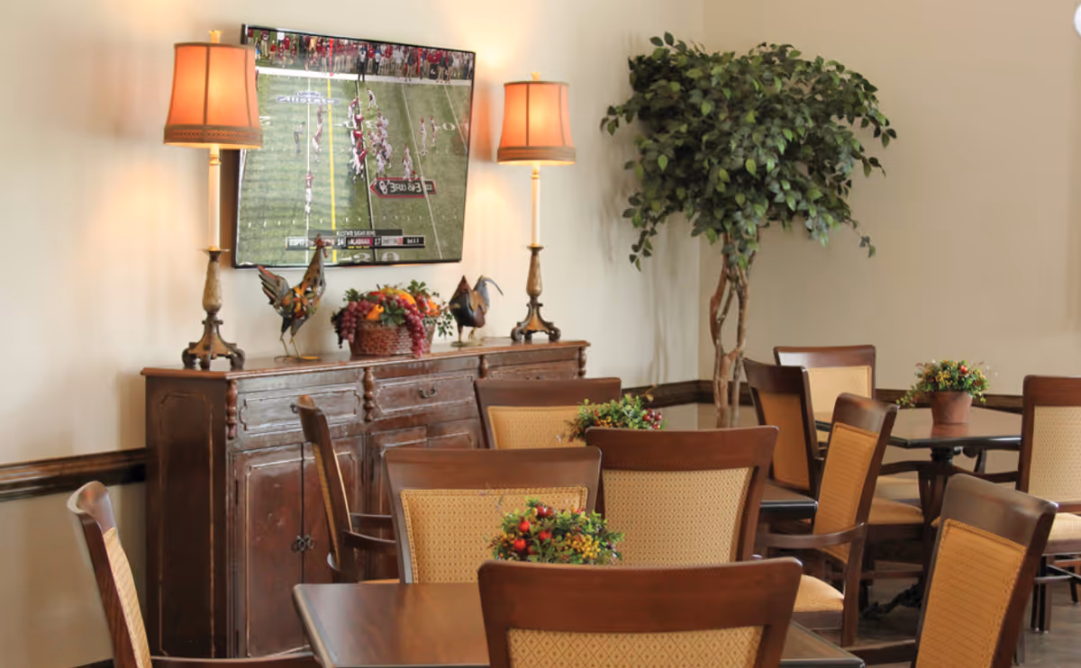 Dining area with wooden tables and upholstered chairs, a sideboard topped with lamps and décor, a wall-mounted TV showing a football game, and a large potted plant.