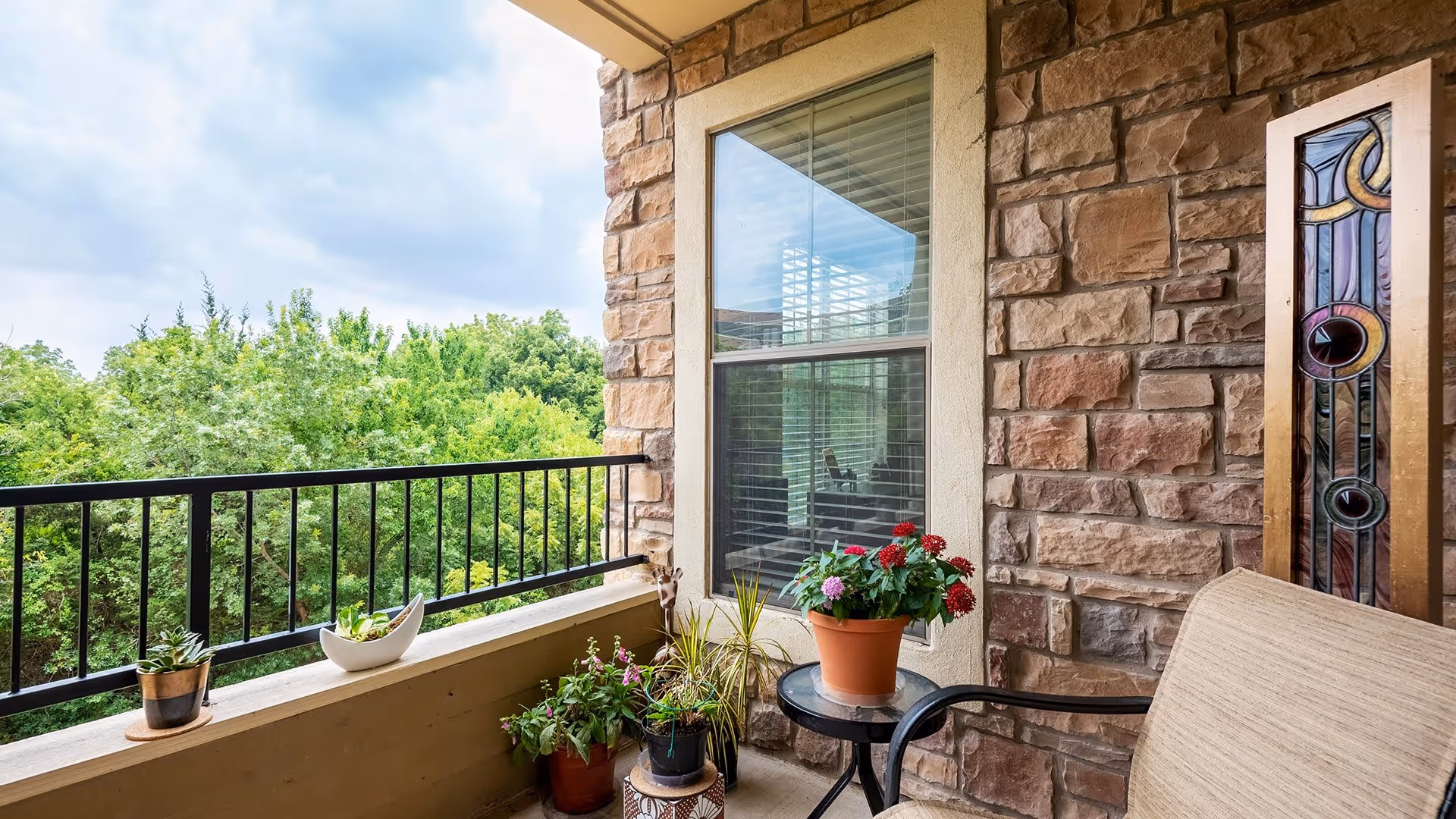 A small outdoor balcony area with a stone wall, a window with blinds, and a black metal railing overlooking green trees. The balcony has several potted plants and flowers placed on the ledge and a small round table next to a beige cushioned chair. The sky is partly cloudy.