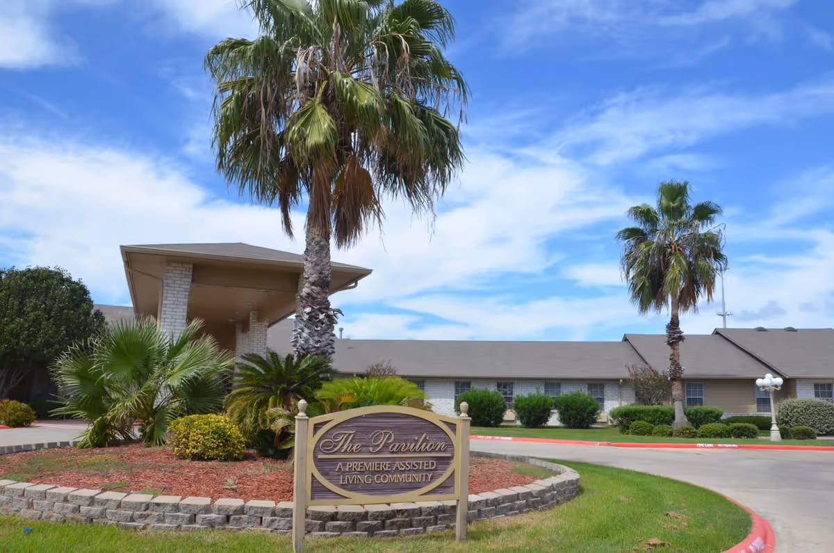 Exterior view of Pavilion Assisted Living facility with a landscaped entrance featuring palm trees, bushes, and a sign that reads 'The Pavilion A Premiere Assisted Living Community' under a partly cloudy blue sky.