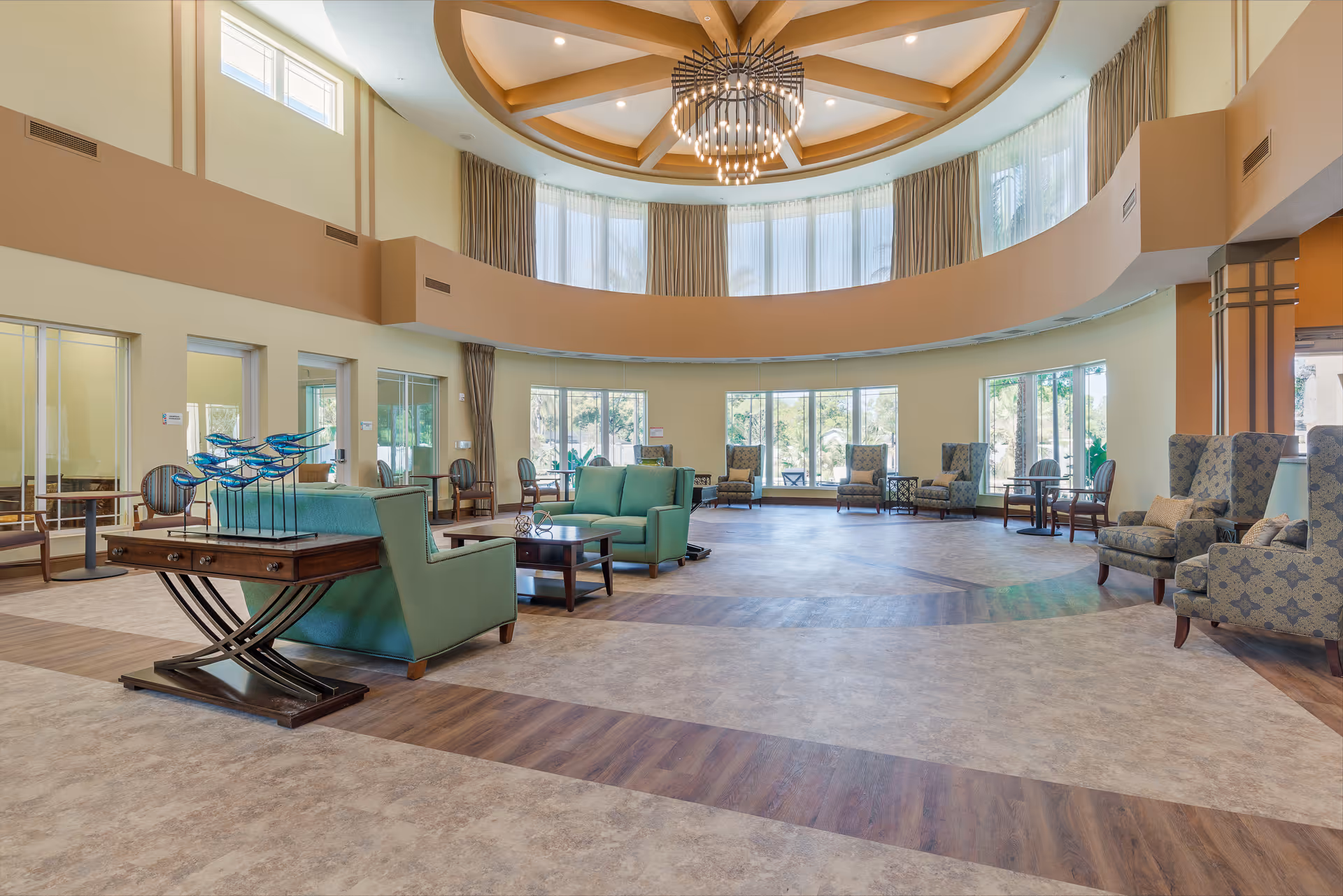 Spacious circular communal lounge with a high coffered ceiling and chandelier, large windows, and grouped sofas and armchairs.
