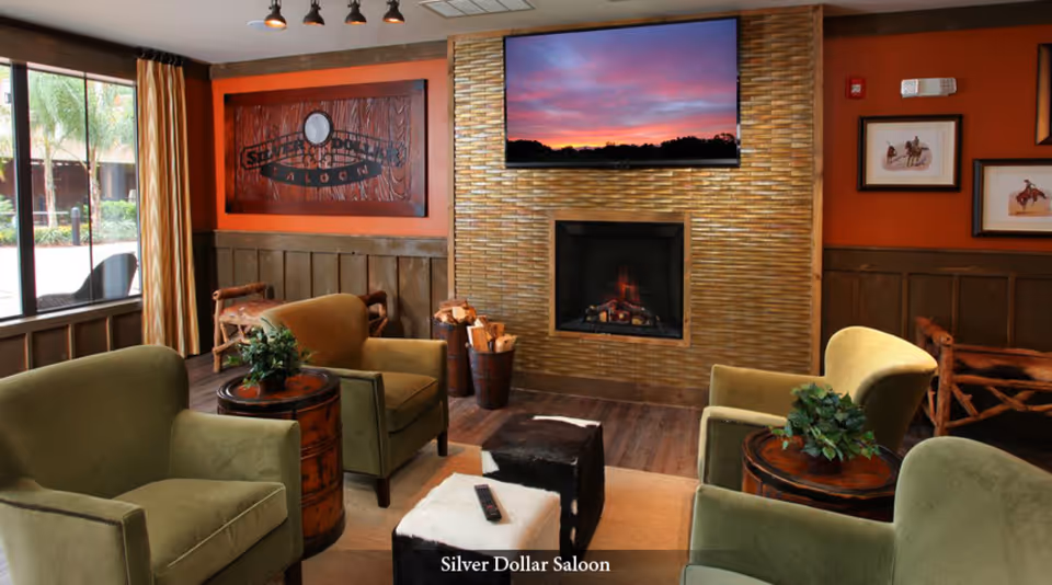 A cozy lounge area with four green armchairs arranged around two small round wooden tables with plants on them. There is a fireplace with a TV mounted above it displaying a sunset scene. The walls are decorated with wooden paneling and framed pictures, and a large window with curtains lets in natural light. A sign on the wall reads 'Silver Dollar Saloon'.