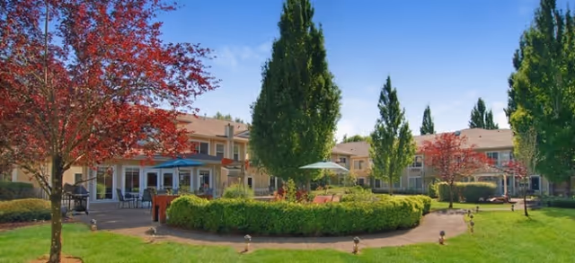 Sunny landscaped courtyard with a circular hedge, trees, outdoor seating, and surrounding two-story senior living buildings.