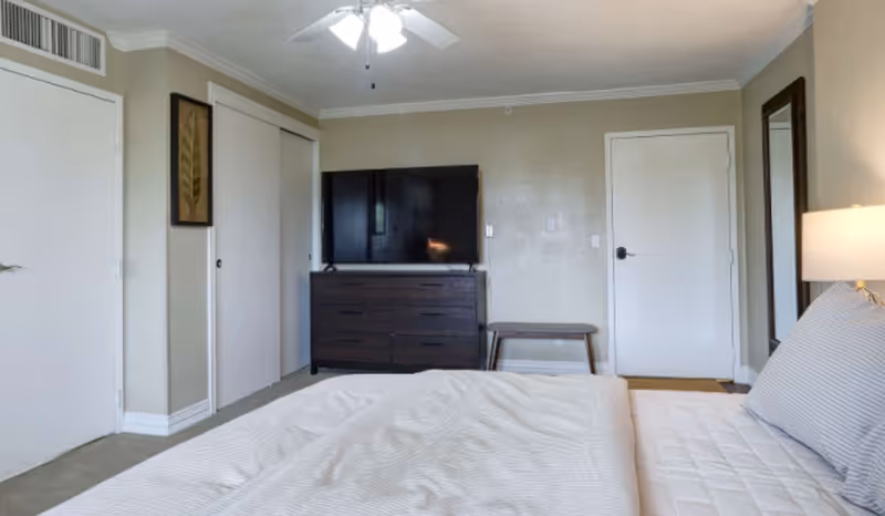 Bedroom with a bed in the foreground, a dresser topped with a TV against the far wall, closet doors, a bench, and a ceiling fan.