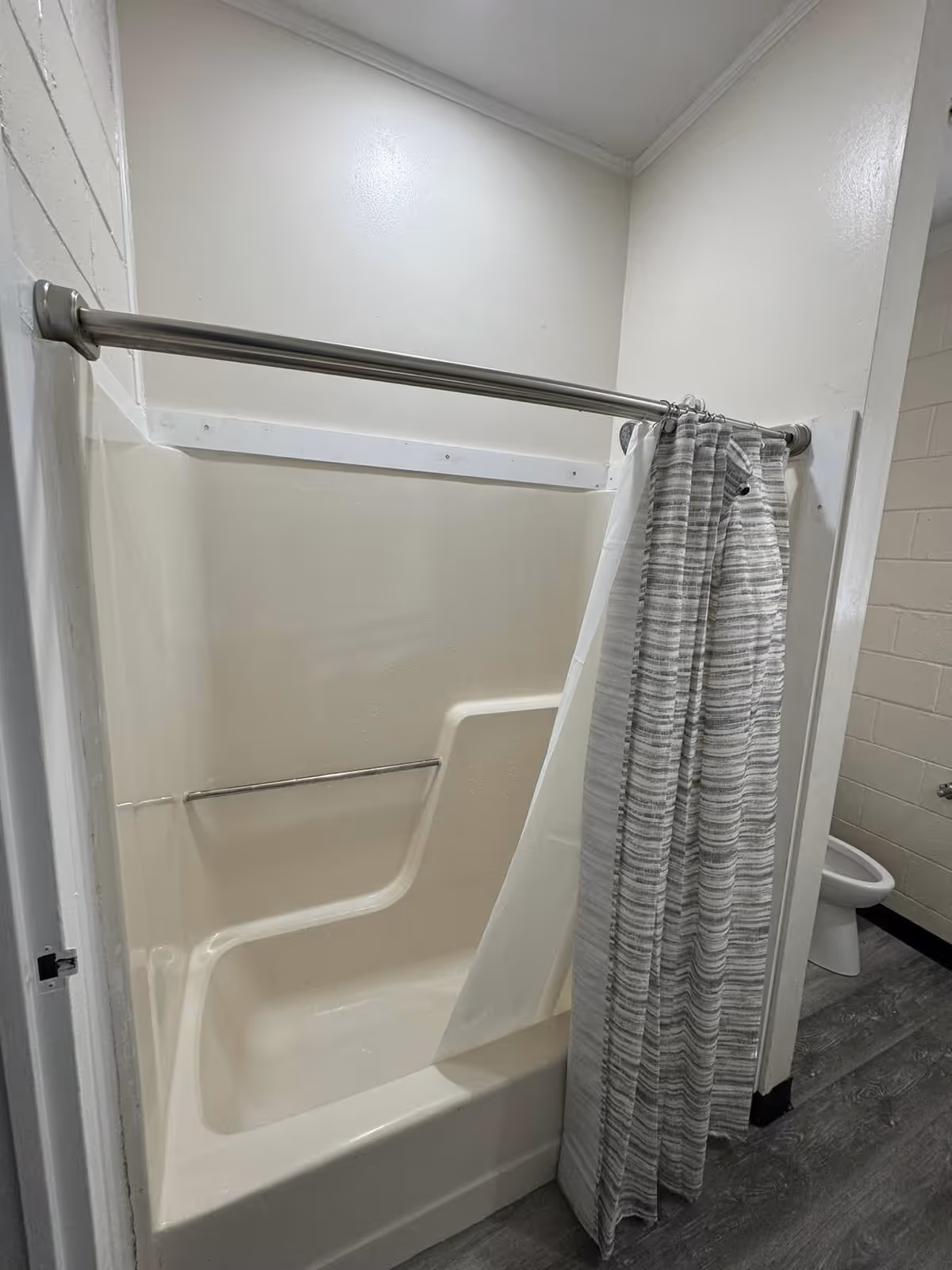A bathroom with a beige bathtub and shower combination featuring a built-in seat. A silver shower curtain rod holds a striped gray and white shower curtain partially pulled to the side. The floor is gray wood-like vinyl, and a white toilet is visible in the adjacent room.