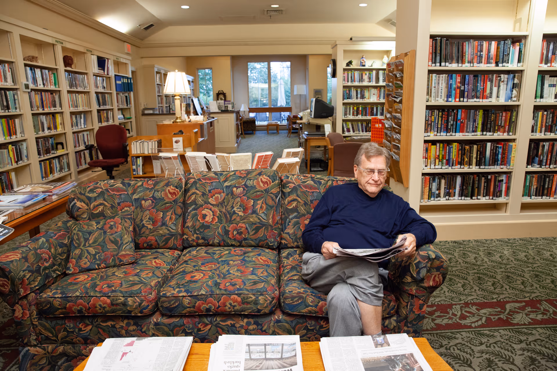 An elderly man wearing glasses and a navy blue sweater sits on a floral-patterned couch in a library or reading room. He is holding a newspaper and appears to be reading. The room is filled with bookshelves stocked with books, a table with newspapers in the foreground, and a desk with a lamp and computer in the background.