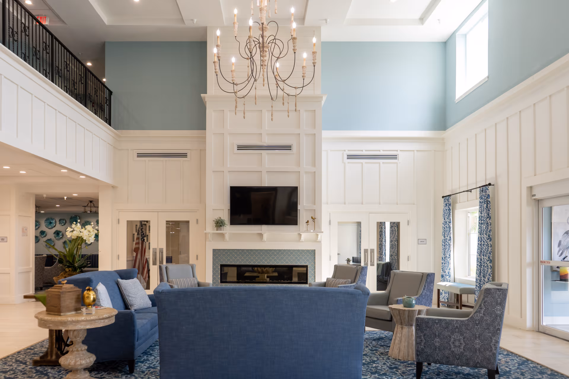 A spacious and bright living room area in Seagrass Village Independent Living featuring high ceilings, a large chandelier, a fireplace with a mounted flat-screen TV above it, and comfortable seating including blue sofas and patterned armchairs arranged around a wooden coffee table. The walls are white with blue accents, and there are large windows letting in natural light.
