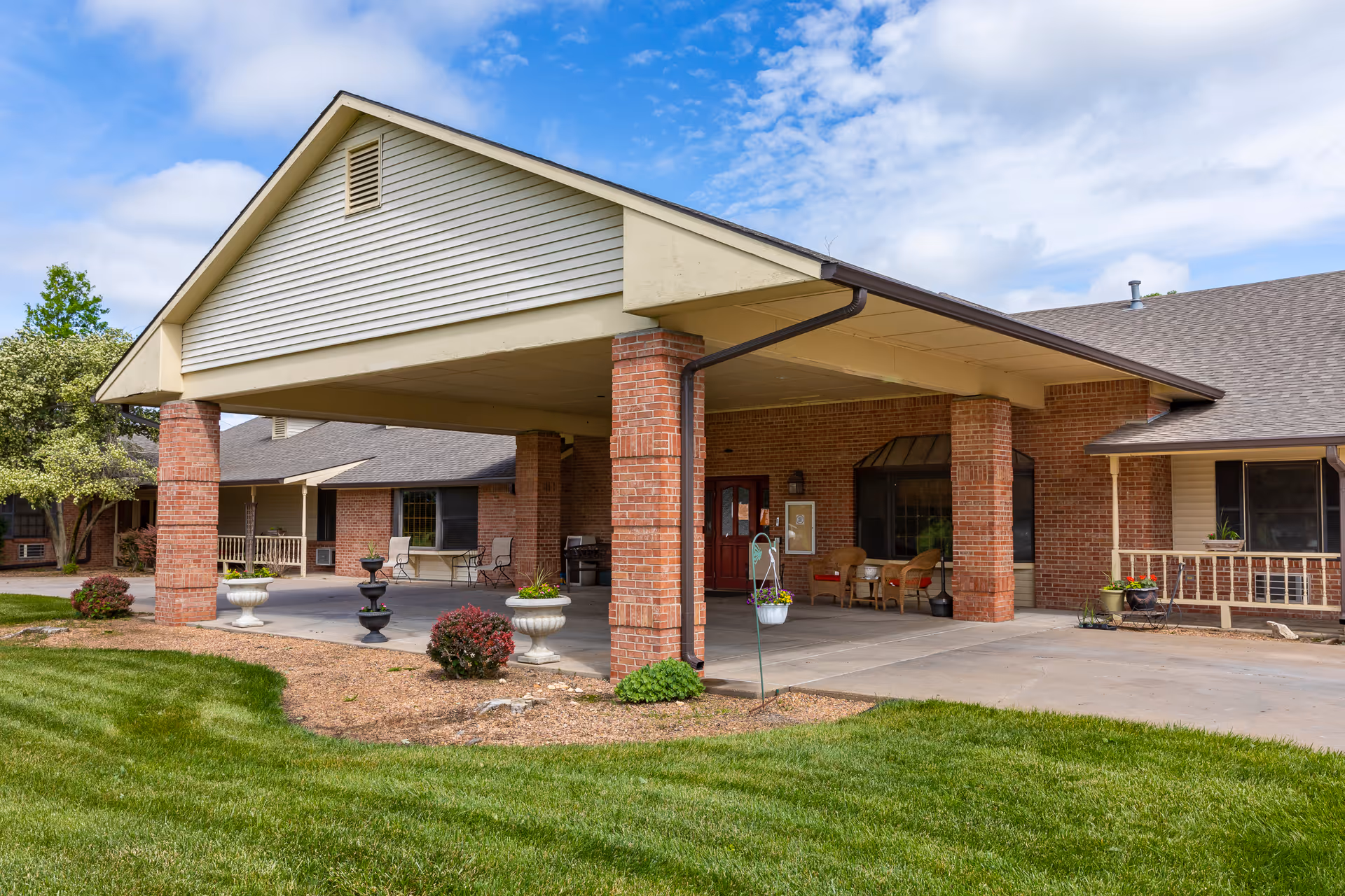 Exterior view of a senior living facility entrance with a covered drop-off area supported by brick columns, surrounded by green grass and landscaping under a partly cloudy sky.