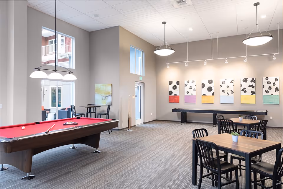 A spacious recreational room with a red pool table on the left, two wooden tables with black chairs on the right, and a shuffleboard table against the far wall. The walls are decorated with colorful abstract paintings, and the room is well-lit with hanging ceiling lights. Large windows and a glass door provide natural light and a view of an outdoor seating area.