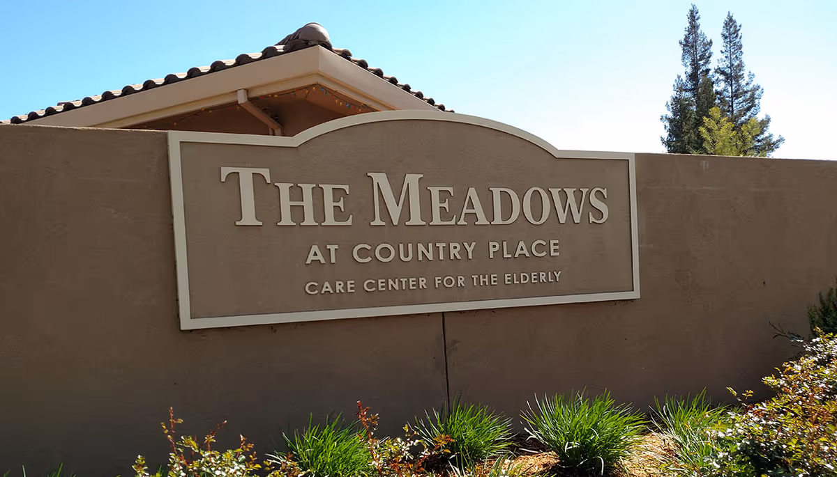 Outdoor sign for The Meadows at Country Place Care Center for the Elderly mounted on a beige wall with a tiled roof structure above it, surrounded by green shrubs and trees under a clear blue sky.