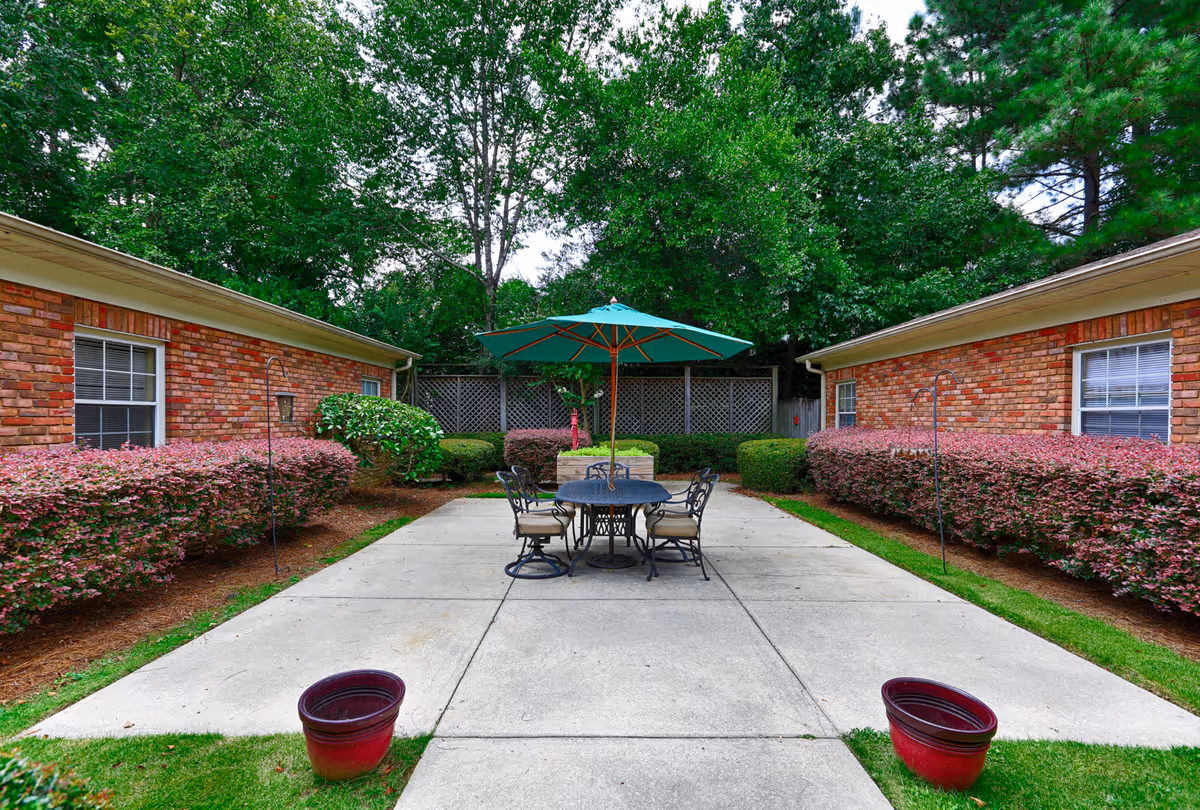Outdoor patio area between two brick buildings with a round table and four chairs under a green umbrella. The patio is surrounded by neatly trimmed bushes and trees in the background.