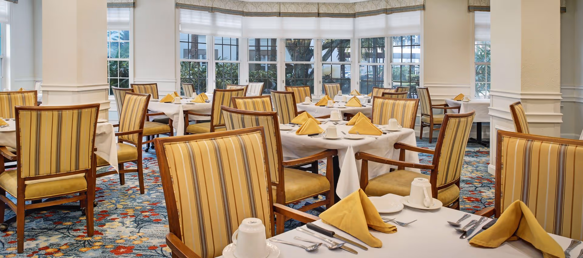 A bright dining room with multiple tables covered in white tablecloths, each set with yellow folded napkins, white cups, plates, and silverware. The chairs have wooden frames with yellow and brown striped upholstery. Large windows with white blinds allow natural light to fill the room, and a colorful patterned carpet covers the floor.