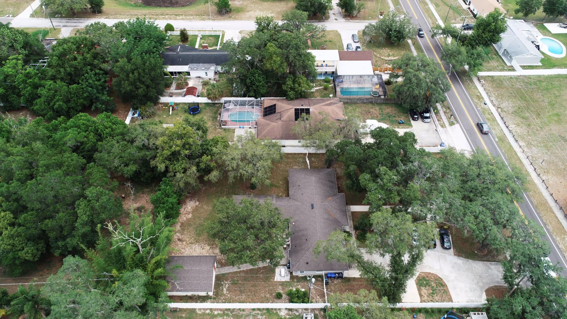 Aerial view of a tree-lined residential property with multiple houses, driveways, and a road at right.