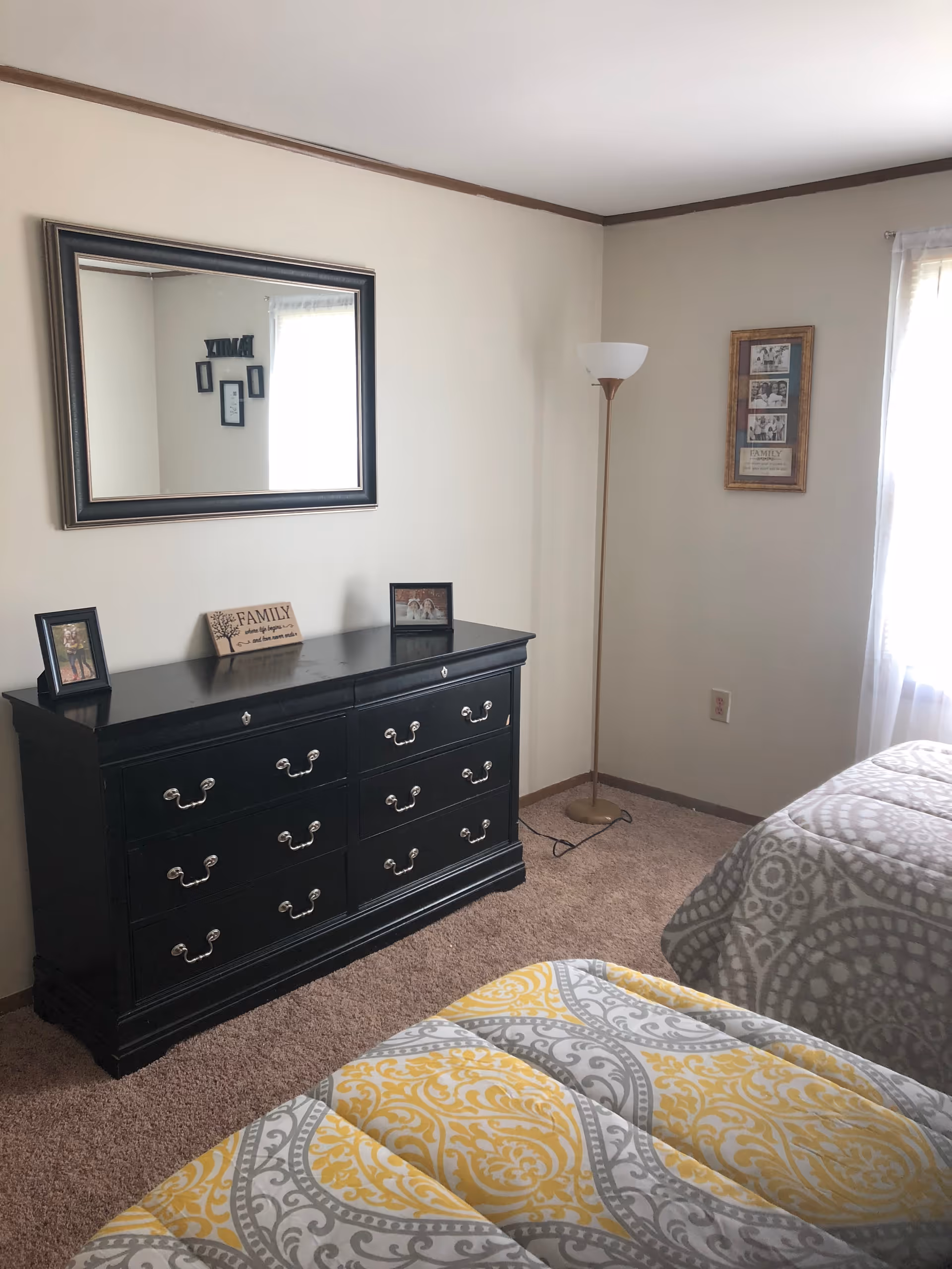A bedroom with two beds covered in patterned yellow and gray bedspreads. There is a black dresser with six drawers against the wall, topped with framed photos and a decorative sign that says FAMILY. Above the dresser is a large rectangular mirror. A tall floor lamp stands in the corner next to a window with sheer white curtains, and a framed picture hangs on the adjacent wall.