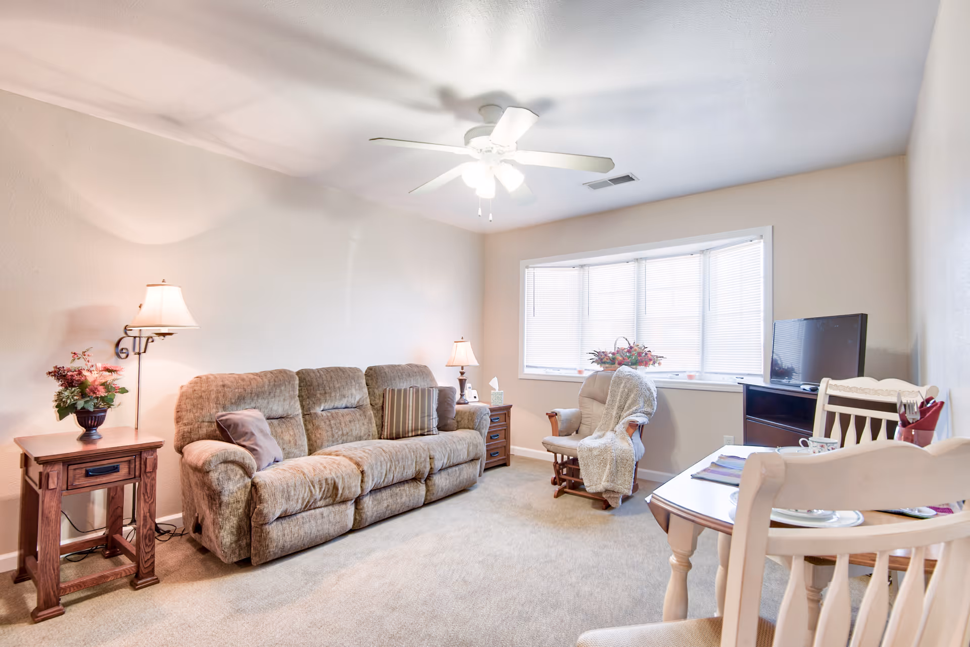 A cozy living room with a beige carpet, a brown upholstered sofa with pillows, a wooden side table with a lamp and a flower vase, a rocking chair with a blanket, a small TV on a stand, and a dining table with chairs. A large window with blinds lets in natural light.
