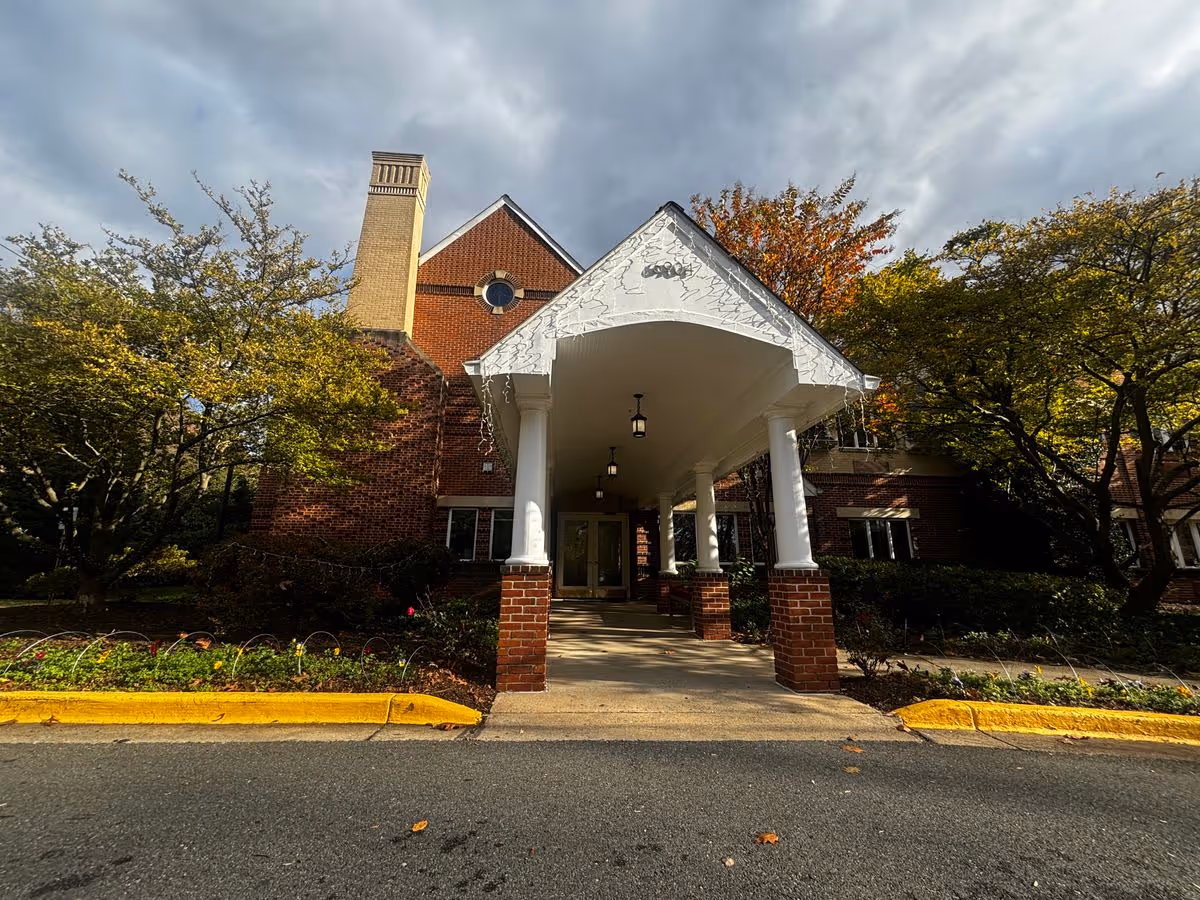 Front entrance of a brick building with a white covered portico supported by columns, flanked by trees and landscaping under a cloudy sky.
